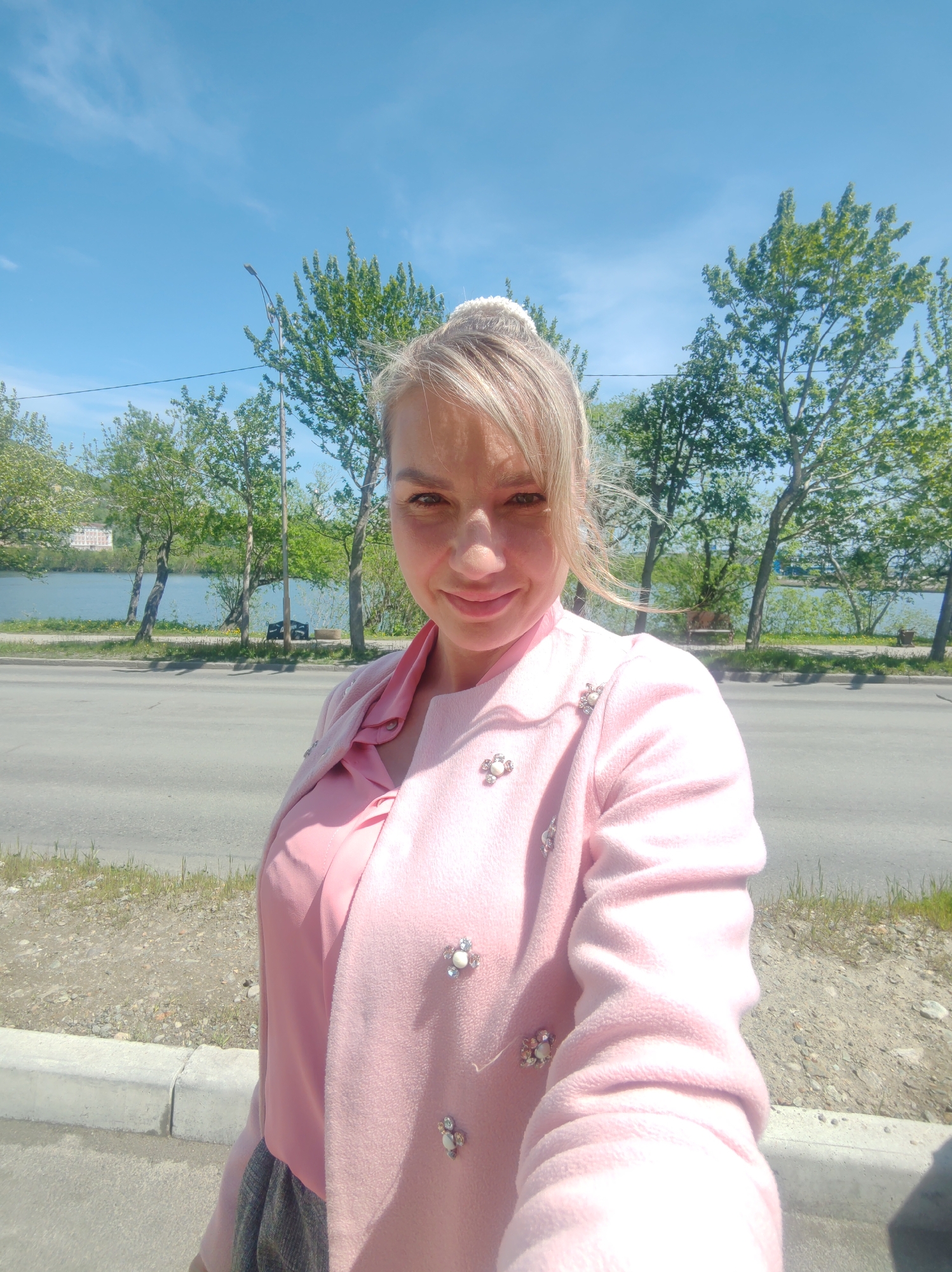 An outdoor, medium shot selfie features a blonde woman with fair skin and light brown eyes, smiling at the camera. She is wearing a pink collared shirt and a pink jacket adorned with floral-shaped embellishments. The background shows a body of water, trees, a road, and a bright blue sky. The woman's hair is pulled back into a bun. The scene appears to be in a park or along a waterfront.