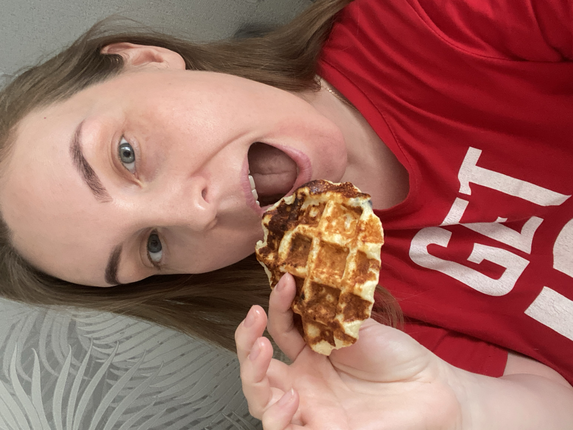 A Caucasian female is depicted in a medium close-up shot, positioned indoors in the city of Noviy, Russia. She holds a browned, square-gridded waffle in her left hand, bringing it toward her open mouth. Her mouth is wide open, revealing the oral cavity. She has light brown hair, light blue eyes, and visible eyebrows. She wears a red t-shirt with white lettering, of which "GET" is partially visible on the left side of the chest. The background consists of a light grey or white wallpaper featuring a repeating pattern of stylized palm or fern leaves.