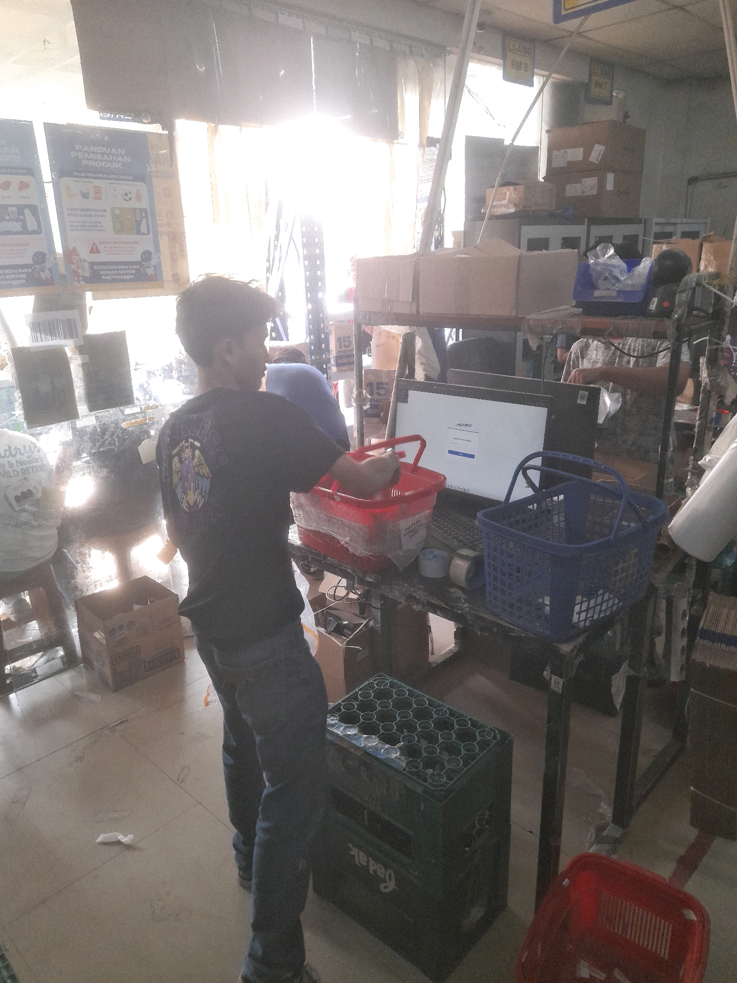The image depicts an indoor scene, likely a retail or storage environment, with a strong ambient light source coming from the left, possibly a window or open doorway, creating a lens flare effect. In the foreground, a young male wearing a dark t-shirt with a graphic design and blue jeans is standing and holding a red plastic basket. He is facing away from the camera, looking towards a cluttered desk area. On the desk, there is a laptop with its screen displaying what appears to be a login interface with the "ASTRO" logo. Several plastic baskets, including a blue one, are also present on the desk, along with rolls of tape and other miscellaneous items. In the background, shelves are stacked with cardboard boxes, and a portion of a blue garment, possibly worn by another person, is visible. The floor is tiled and shows some signs of wear and tear, with a few white paper-like objects scattered around. The overall impression is of a busy, somewhat disorganized workspace, possibly within a small business or warehouse setting. Signs in Indonesian, such as "PANDUAN PEMISAHAN PRODUK" (Product Separation Guide) and "BELI DENGAN PENUH KEYAKINAN" (Buy with Full Confidence), are visible on the wall to the left, suggesting the location could be a store. Given the context of the "ASTRO" logo on the laptop screen, which is a telecommunications company in Malaysia, and the presence of Indonesian language signs, the location could plausibly be a business operating in or associated with Bekasi, Indonesia.