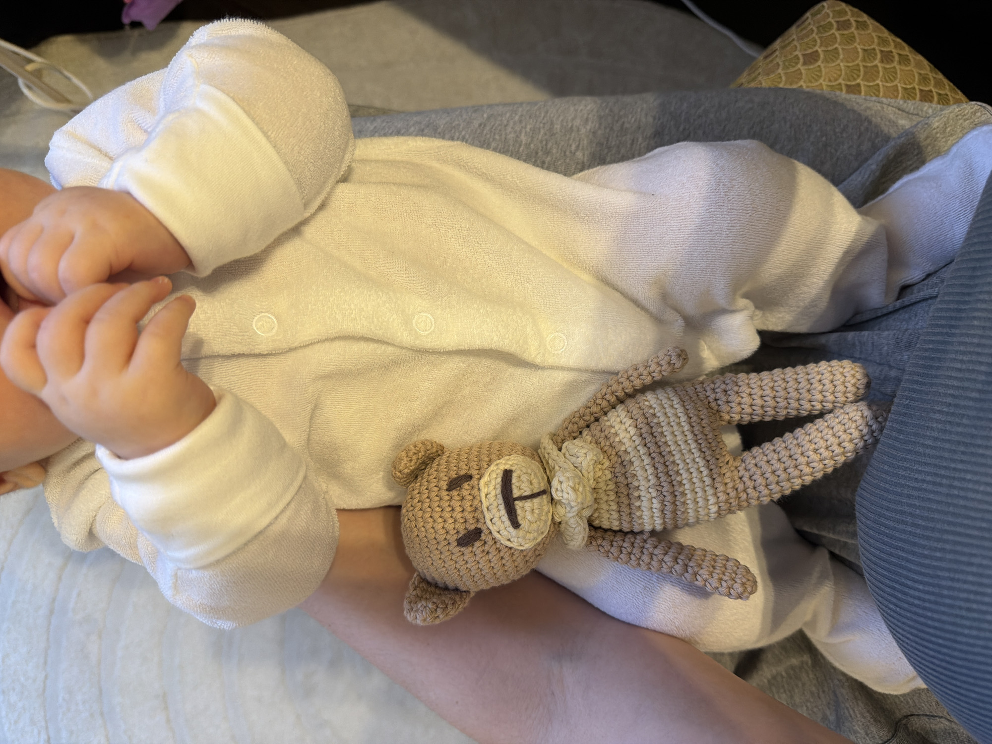A baby in a white onesie with fold-over mittens lies on a person's lap. The baby is holding their fingers to their mouth. A crocheted bear is lying next to the baby. The person's lap is covered with gray and blue fabric. The location is unknown, and Khimki, Russia cannot be confirmed.