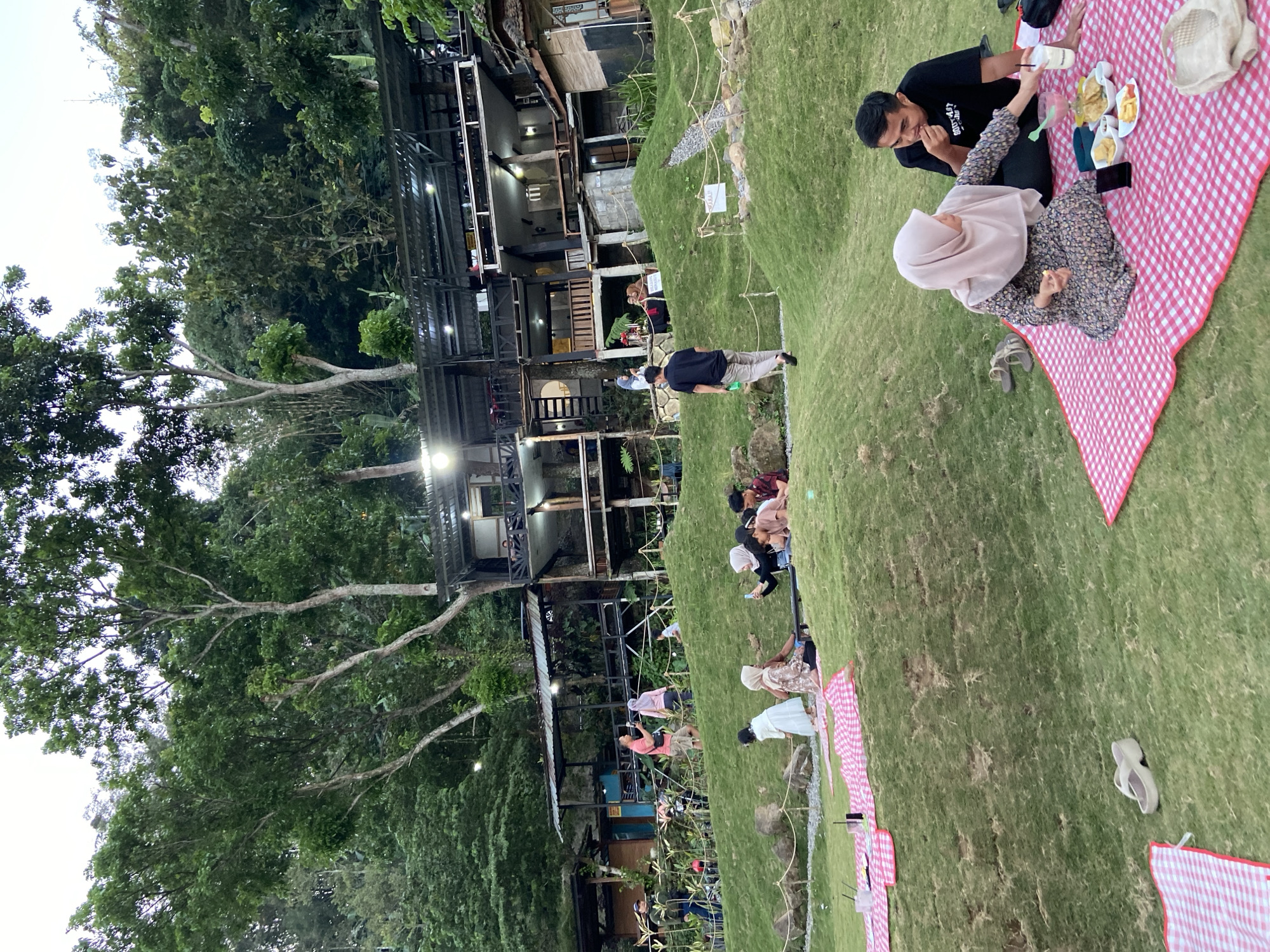 The image depicts an outdoor gathering at a tiered, hillside venue in Kesiman, Indonesia. The scene features a cluster of multi-level buildings with open-air balconies and roofs, nestled amongst dense green foliage. Several groups of people are scattered across the grassy slopes, engaging in various activities. In the foreground, a couple is having a picnic on a red and white checkered blanket, with food and drinks laid out. Other individuals are seen sitting and walking on the grass. Some appear to be taking pictures with their phones. Red and white checkered blankets are spread in multiple locations on the lawn, suggesting a communal or recreational space. The environment is lush and green, with trees and vegetation dominating the upper portions of the image.