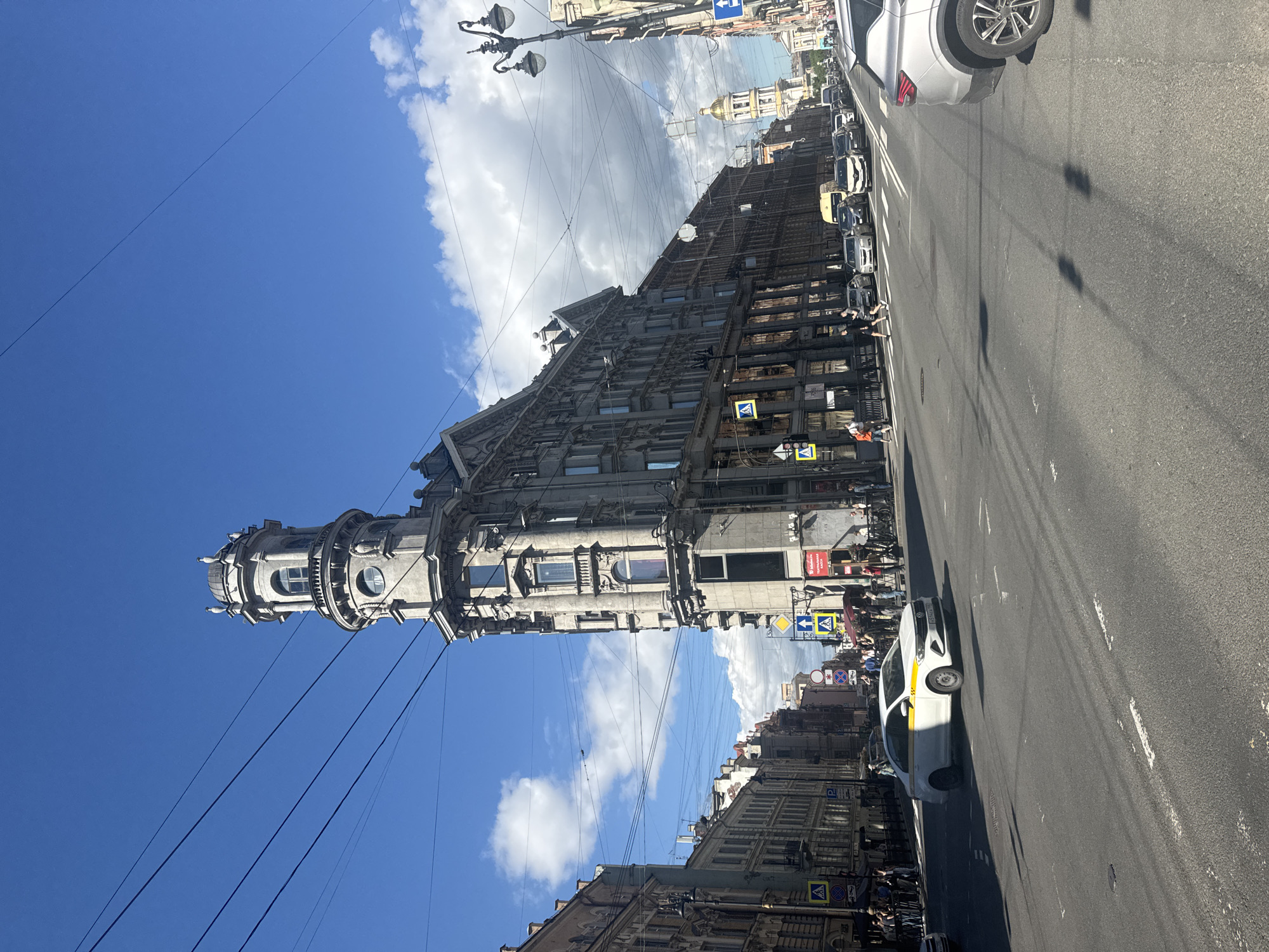 This is a street-level, eye-level, daytime, medium-shot photograph taken from a street intersection in Saint Petersburg, Russia. The prominent architectural feature in the center is the Singer Building, also known as the Dom Knigi, a striking Art Nouveau building with a distinctive rounded corner tower topped by a glass globe and spire. The building's façade is a light grey stone, with numerous windows and decorative elements.
To the left of the Singer Building, other historic multi-story buildings line the street, with detailed stone facades and numerous windows. To the right, the street continues, lined with similar architectural styles, leading to a distant view of a bell tower with a golden dome.
The street is paved with asphalt and marked with white lane lines and pedestrian crossings. Several cars are visible on the road, including a white taxi with a yellow stripe. People are walking on the sidewalks and crossing the street. The sky is bright blue with scattered white clouds, and overhead, a complex network of telephone or electrical wires is visible. The overall scene depicts a busy urban environment on a clear day.