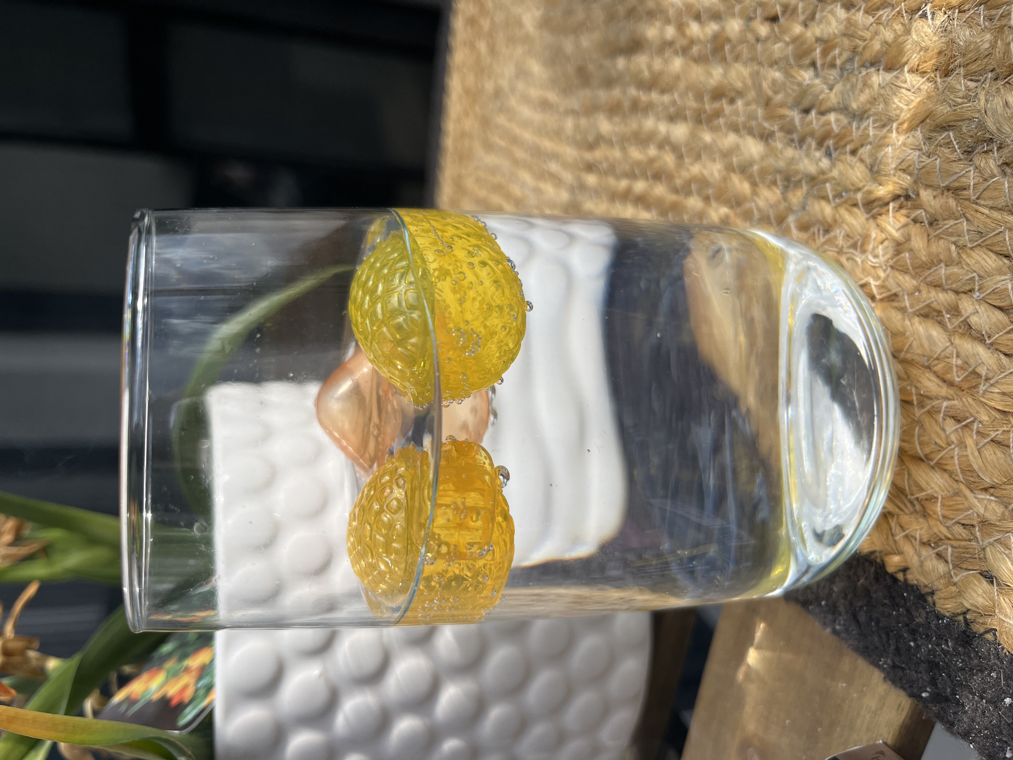 The image shows a clear glass filled with water, lying on its side. Inside the glass are two yellow, textured balls with small air bubbles clinging to their surfaces. A small, light-brown object is also visible within the water. The glass rests on a surface, partially covered by a woven, tan-colored material, and adjacent to a white, textured object, possibly a planter. A plant with green and yellowish foliage is visible on the left. The background is blurred, showing a dark-colored area above and a lighter, white area below.
There is not enough information to determine the specific location in Varna, Bulgaria.