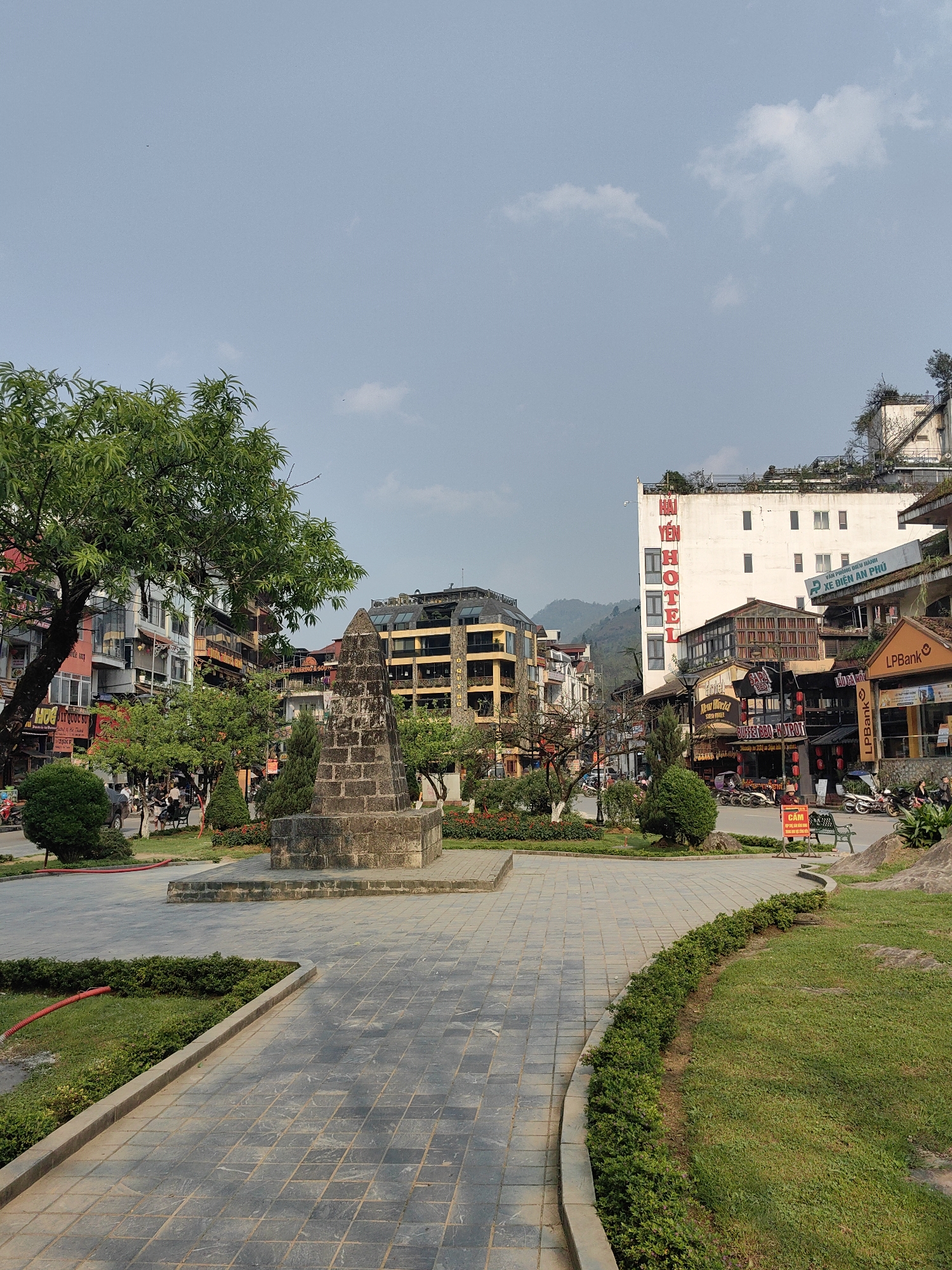 This image depicts a public square or park area in the city of Sa Pa, Vietnam, on a clear day with a light blue sky and scattered white clouds. In the foreground and center-left, a weathered dark grey stone monument, constructed from stacked rectangular blocks, stands on a raised platform. Paved grey tiled pathways curve through the scene, bordered by neatly maintained green grass and low green hedges. A large leafy green tree occupies the left foreground, partially obscuring multi-story buildings. A red coiled hose lies on the grass near the left pathway.
Beyond the park, a two-lane paved street is lined with numerous commercial buildings. On the left side of the street, various shops and restaurants are visible, with signage primarily in Vietnamese. A grey SUV is partially visible parked on this side. On the right side of the street, a prominent white multi-story building displays large vertical red text "HAI YEN HOTEL." Below it, other businesses include an "LPBank" branch, "NEW WORLD," and "SAPA HOTPOT BBQ-HOTPOT," with associated signage and parked motorbikes. Several individuals are visible on the sidewalks and near the parked vehicles. A green park bench and a red sign with Vietnamese text "CẤM" (FORBIDDEN) are positioned on the right side of the main road. Distant mountains are visible in the background, behind the urban structures.