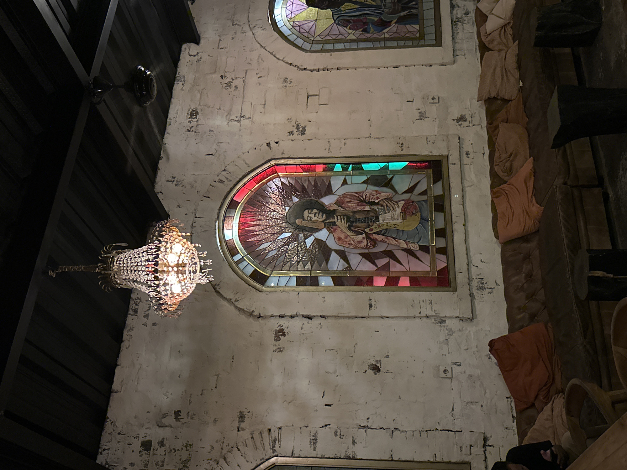 The image captures a dimly lit, ornately decorated interior, likely a restaurant or bar, characterized by a unique blend of rustic and artistic elements. The walls are made of distressed white-washed brick or stone, showing texture and age, while the ceiling or upper structure is dark and possibly slatted.
The most striking features are two arched stained glass windows. The lower, more prominent window depicts a stylized figure, strongly resembling Jimi Hendrix, playing an electric guitar. This artwork is vibrant with reds, oranges, and greens, featuring sunburst patterns emanating from behind the figure. A second, partially visible stained glass window above it also displays a figural design in a similar artistic style.
A large, crystal-beaded chandelier hangs from the ceiling on the left side, emitting a warm, inviting glow that illuminates its intricate details. To the right, cushioned seating or banquettes are visible, adorned with draped fabrics or pillows in earthy tones like tan and orange-brown, suggesting a comfortable lounge area. Dark, robust furniture elements are incorporated into this seating.
Additional details include a dark-colored fixture, possibly a ceiling fan or light, mounted on the dark ceiling in the upper left, and a white electrical outlet on the white wall. No people are present in the scene, and no specific activity is taking place. The overall ambiance, created by the dim lighting, the warm glow of the chandelier, and the distinctive stained glass art, suggests an evening setting designed for dining, socializing, or enjoying live music, given the prominent musician depicted. No visible text is present.
