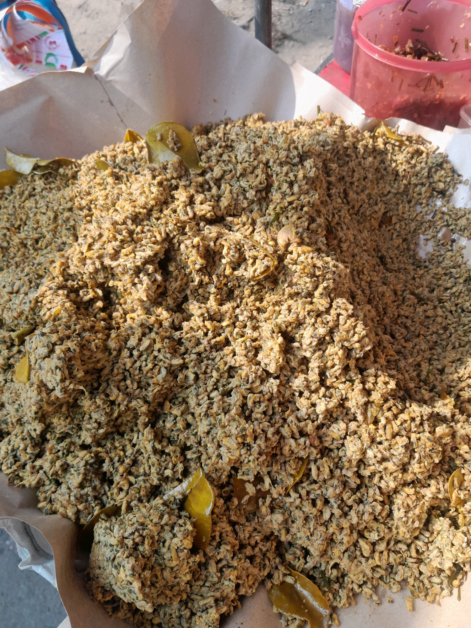 The image shows a close-up, overhead view of a pile of food, likely a culinary dish, placed on brown paper packaging. The food appears granular and is a light brown to yellowish-brown color with some darker specks and occasional green leaf-like fragments mixed in. The texture suggests it might be small grains, seeds, or possibly finely chopped ingredients.
In the background, a translucent pink plastic container with a red lid is visible, containing a reddish mixture with some dark green and red elements. To the left and slightly behind the food pile, a colorful plastic bag with stripes of blue, white, orange, and red is partially visible. The ground surface appears to be sandy or dusty. The lighting suggests an outdoor environment with sunlight casting shadows. The overall scene suggests a food stall or market setting, potentially in Jogosatru, Indonesia, given the context provided. There are no people directly visible in the frame, and no specific actions or interactions are depicted, other than the passive presentation of the food.