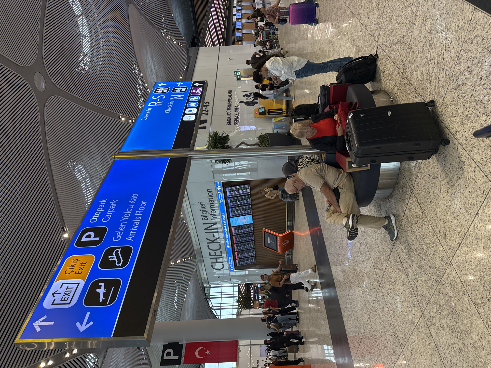 This image depicts the interior of a modern airport terminal in Arnavutköy, Turkey. The scene is brightly lit with a high, contemporary ceiling structure featuring linear metallic elements. In the foreground, several individuals are seated or standing, some with luggage. Two individuals are seated on a curved bench, one man looking down at his phone and a woman looking forward. In front of them are several pieces of luggage, including a large black suitcase. To the right, a woman in blue jeans stands looking at her phone. In the background, a large departures/arrivals board displays flight information. A prominent blue overhead directional sign indicates "CHECK-IN Bilgileri Information" and displays various directions and icons, including those for "Otopark," "Carpark," and "Arrivals Floor." Another blue sign above it indicates "Check-in R-S" and "Check-in N." Other elements visible include a Turkish flag hanging from a pillar and various kiosks and airport facilities. The floor is a polished, speckled granite material. The overall atmosphere suggests a busy travel hub.