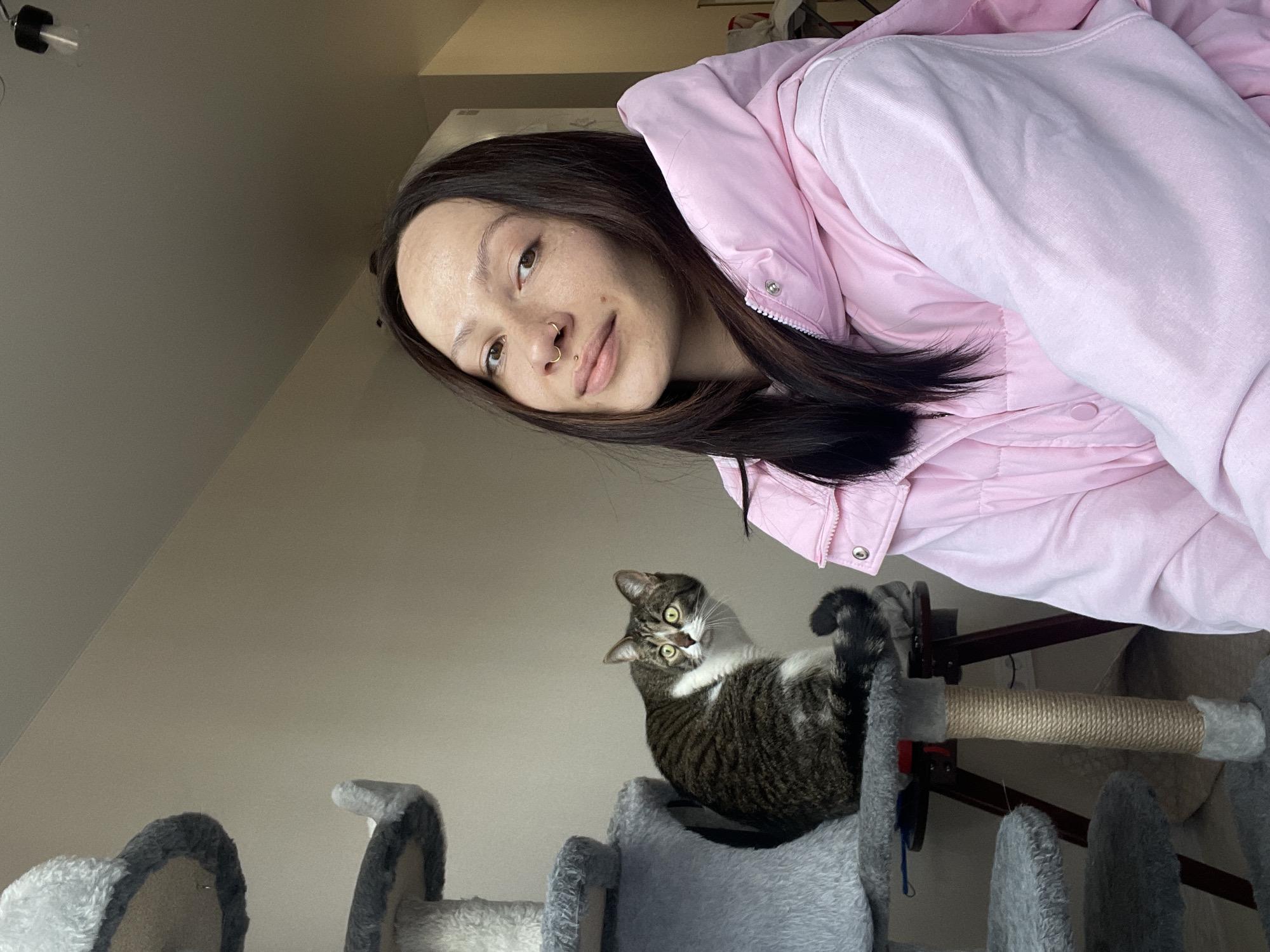The image, captured in Izhevsk, Russia, presents a tilted, low-angle shot of a woman and a cat in an indoor home setting. The woman, positioned in the upper right, has long dark hair, light eyes, and a small piercing on her nose. She wears a light pink puffy jacket over a matching light pink hoodie and gazes towards the camera with a gentle smile.
Below her and to the left, a tabby cat with distinctive brown and black stripes and white markings on its chest and paws is perched on a grey, multi-level cat tree. The cat's eyes, wide and green-yellow, are fixed directly on the camera, conveying an alert and curious expression. Its tail is curled around its body, and one paw is slightly lifted. The cat tree features scratching posts wrapped in rope.
The background is a simple, light-colored wall, indicating an uncluttered domestic environment. The lighting suggests it is daytime. The primary activity is posing for a photograph. No visible text is present in the image. The scene offers a casual glimpse into the companionship between the woman and her feline pet.