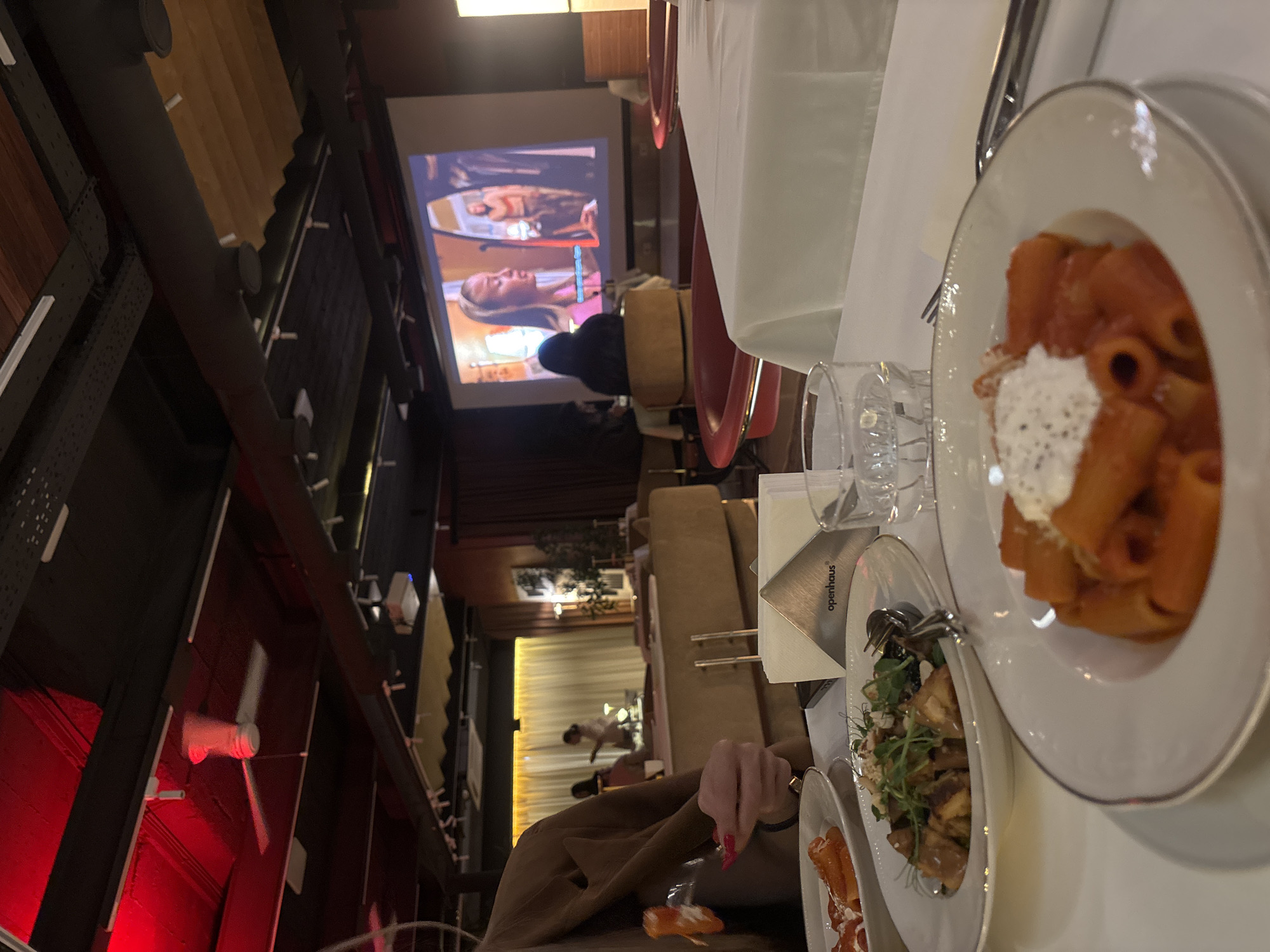 The image captures a dining scene within a restaurant, where a person is actively eating with a fork. Food, including pasta with tomato sauce and ricotta cheese, is visible on the table. The restaurant appears to be playing a movie on a projection screen in the background, adding to the ambiance. Architectural elements like ceiling fans and the building's structural support system are discernible, suggesting a rustic or industrial-chic design. The name "Openhaus" is visible on a piece of paper. The location is Yerevan, Armenia.