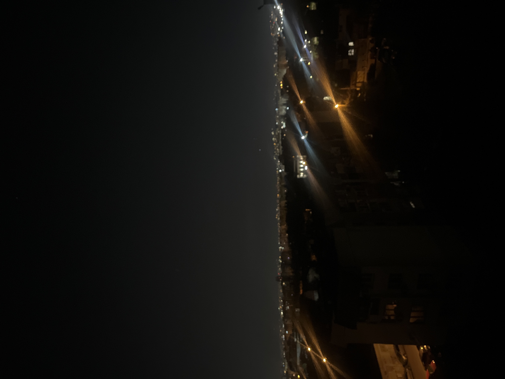 The image presents a panoramic night view of a city, initially oriented in a portrait aspect but depicting a horizontal cityscape. The upper portion of the frame is dominated by a vast, dark sky, subtly illuminated by a hazy glow originating from the city lights below, characteristic of urban light pollution.
The lower half of the image showcases a continuous expanse of artificial lights, stretching across the horizon, indicating a sprawling urban environment with buildings and streets. These lights vary in intensity and color, primarily appearing as white and amber points and streaks of illumination against the profound darkness. Some lights are distinct, possibly streetlights, while others are blurred, suggesting a wide shot or perhaps motion blur. The silhouettes of buildings are faintly discernible on the right side, defined by the interior or exterior lights they emit.
The scene captures a static moment of a city at night. No people or specific activities are visible within the frame. The time of day is unequivocally nighttime. There are no prominent weather details other than the atmospheric haze reflecting the city's glow. Based on the provided information, the location depicted is Sofia, Bulgaria. No visible text appears in the image.