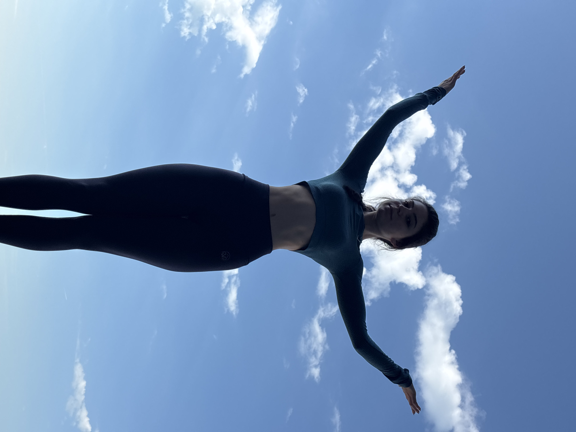 An outdoor, full-body shot captured from a low-angle perspective, showcases a young woman silhouetted against a vibrant blue sky dotted with scattered white cumulus clouds. The woman is positioned centrally, with her body straight and her arms extended horizontally or slightly upwards to her sides, evoking a sense of flight, balance, or a graceful dance/yoga pose.
She is wearing dark, possibly teal or deep green, athletic attire, comprising a long-sleeved crop top that reveals a portion of her midriff, and dark leggings. The strong backlighting from the sun creates a distinct silhouette of her form, highlighting her posture. Despite the silhouette, a subtle hint of her facial features is visible, suggesting a peaceful or smiling expression. A small, circular logo or design is discernible on the upper left thigh of her leggings.
The scene depicts a clear and bright daytime, likely mid-day, given the intensity of the light and the high contrast. The weather appears pleasant with fluffy clouds scattered across the expansive sky. The overall impression is one of serenity and dynamic movement against the open air, consistent with an outdoor activity in a location like Varna, Bulgaria. There is no explicit text visible, only the described logo.