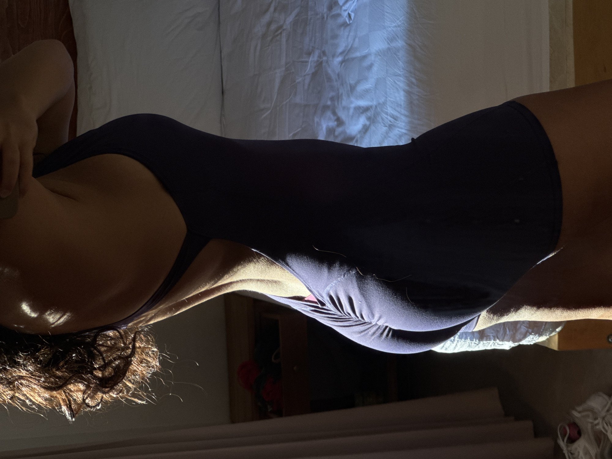 A woman wearing a dark-colored bodysuit is lying on a white bed. The lighting is dramatic, with strong contrasts between light and shadow. The location is possibly North Kuta, Indonesia.