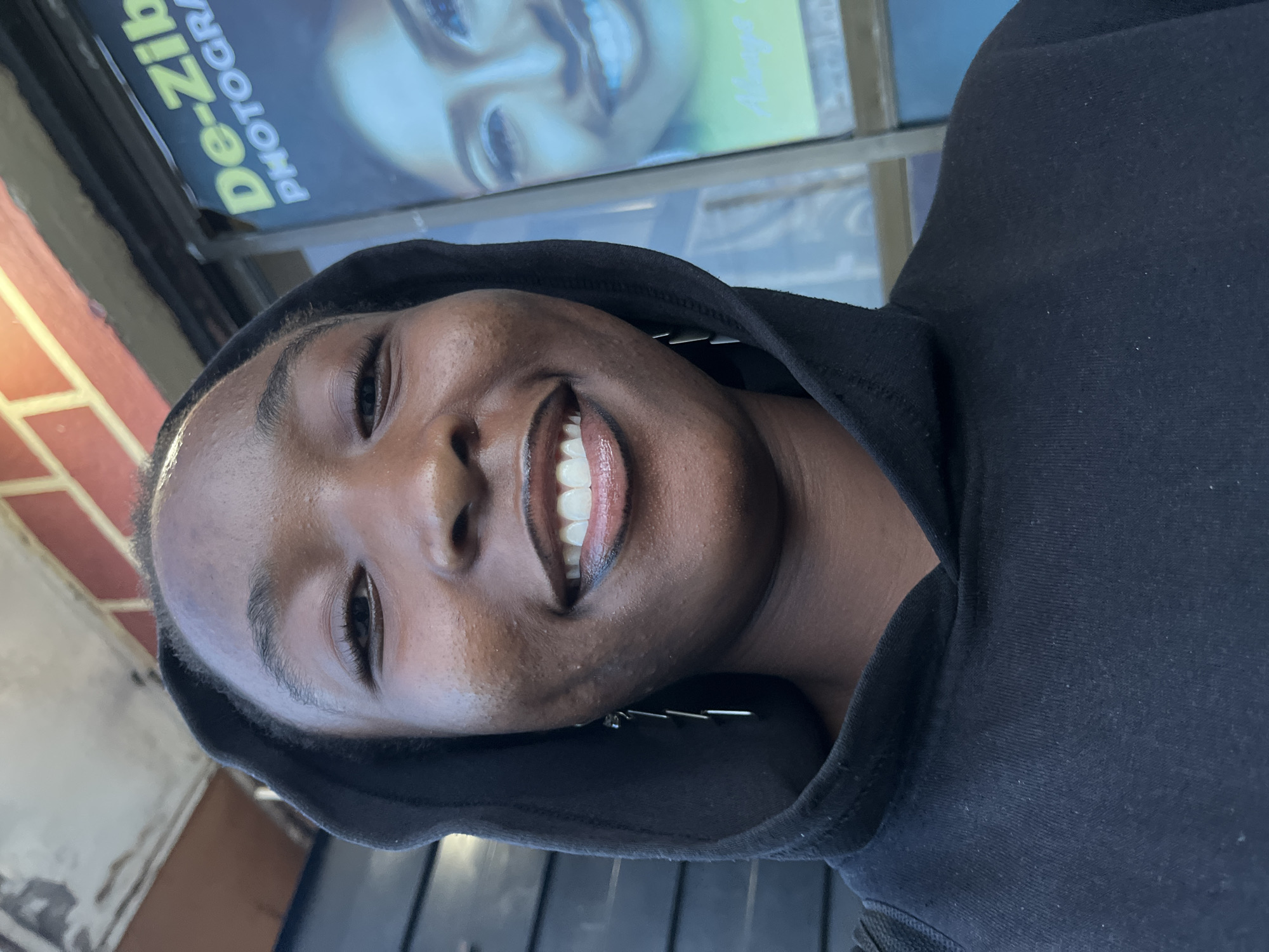 A young Black individual is captured in a close-up, selfie-style portrait, smiling directly at the camera. They possess a dark complexion, displaying natural skin texture with visible pores, particularly on the cheeks. Their dark brown eyes are crinkled at the corners due to a wide, genuine smile that reveals their upper teeth. Full lips are outlined with a dark liner and filled with a lighter, glossy shade. The individual wears a black hooded sweatshirt (hoodie), the hood pulled up around their head. A long, multi-segment silver-colored dangle earring is visible on their left ear (viewer's right).
The background indicates an urban setting, likely a storefront in the city of Unknown, Unknown. To the upper left, a weathered, light-colored concrete or stucco overhang is visible above a dark, possibly wooden or metal wall paneling. On the upper right, a section of a red brick wall is present. Dominating the right side of the background is a large, glass-encased vertical poster or sign. The text "De-Zib" is prominently displayed in bright yellow-green letters, with "PHOTOGRA" (likely "PHOTOGRAPHY") beneath it in lighter lettering, suggesting a business name. The poster features a digitally stylized image of another smiling Black woman, rendered in cool blue and green tones, emphasizing her eyes and teeth. A lighter text tagline is visible at the bottom of the poster.