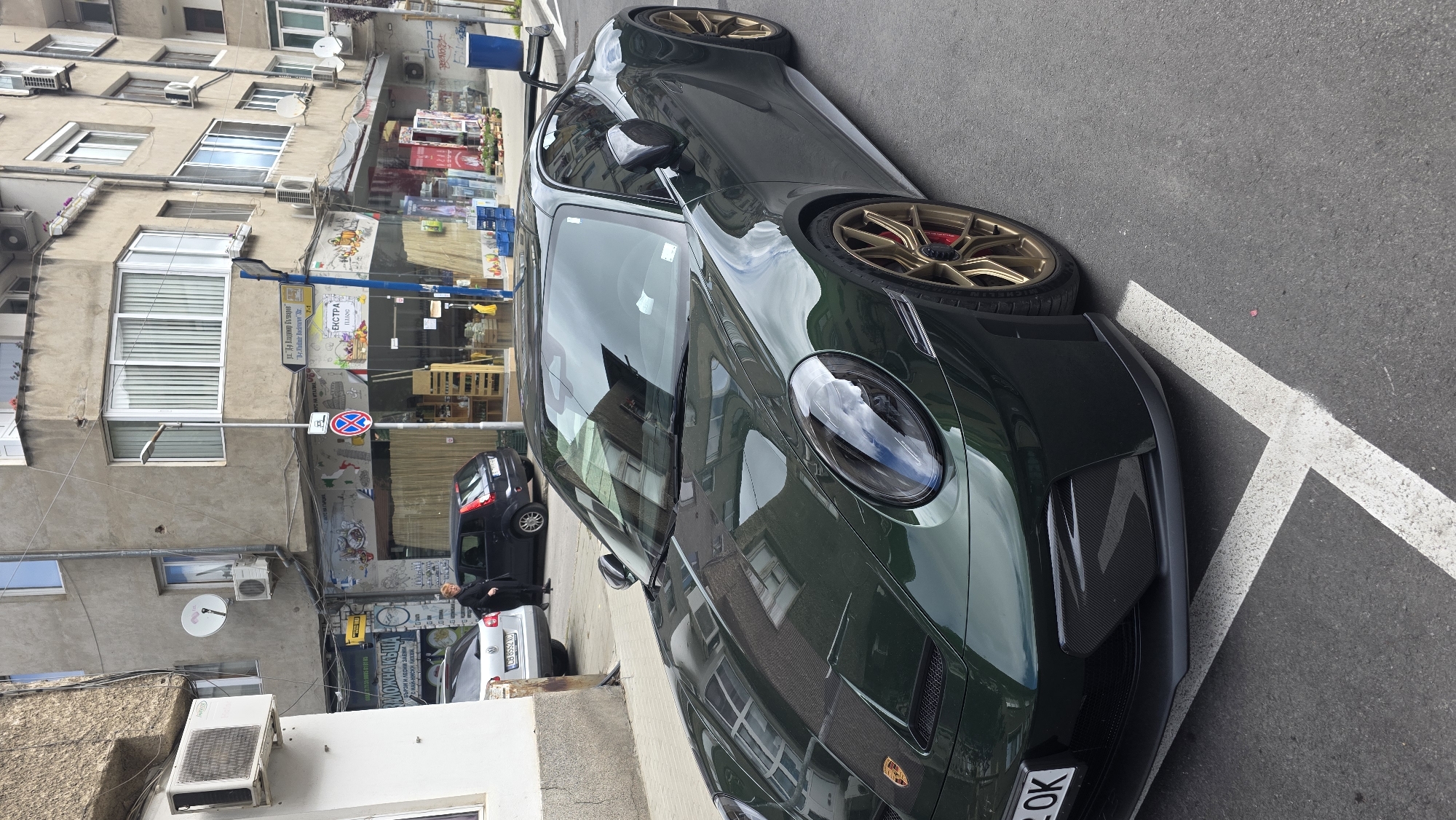 Here's a forensic description of the image:
The image shows a dark green Porsche 911 GT3 parked on the side of a street in Blagoevgrad, Bulgaria. The car has bronze-colored wheels. The street is lined with buildings of a residential and commercial nature. A small black car is visible behind the Porsche, along with a silver sedan. A woman in dark clothing is seen standing near the silver sedan. Street signs are visible, including one indicating "Vladimir Bachvarov Str." in Bulgarian. The environment appears urban, with signs of age and wear on the buildings. A white directional marking is painted on the street next to the car.