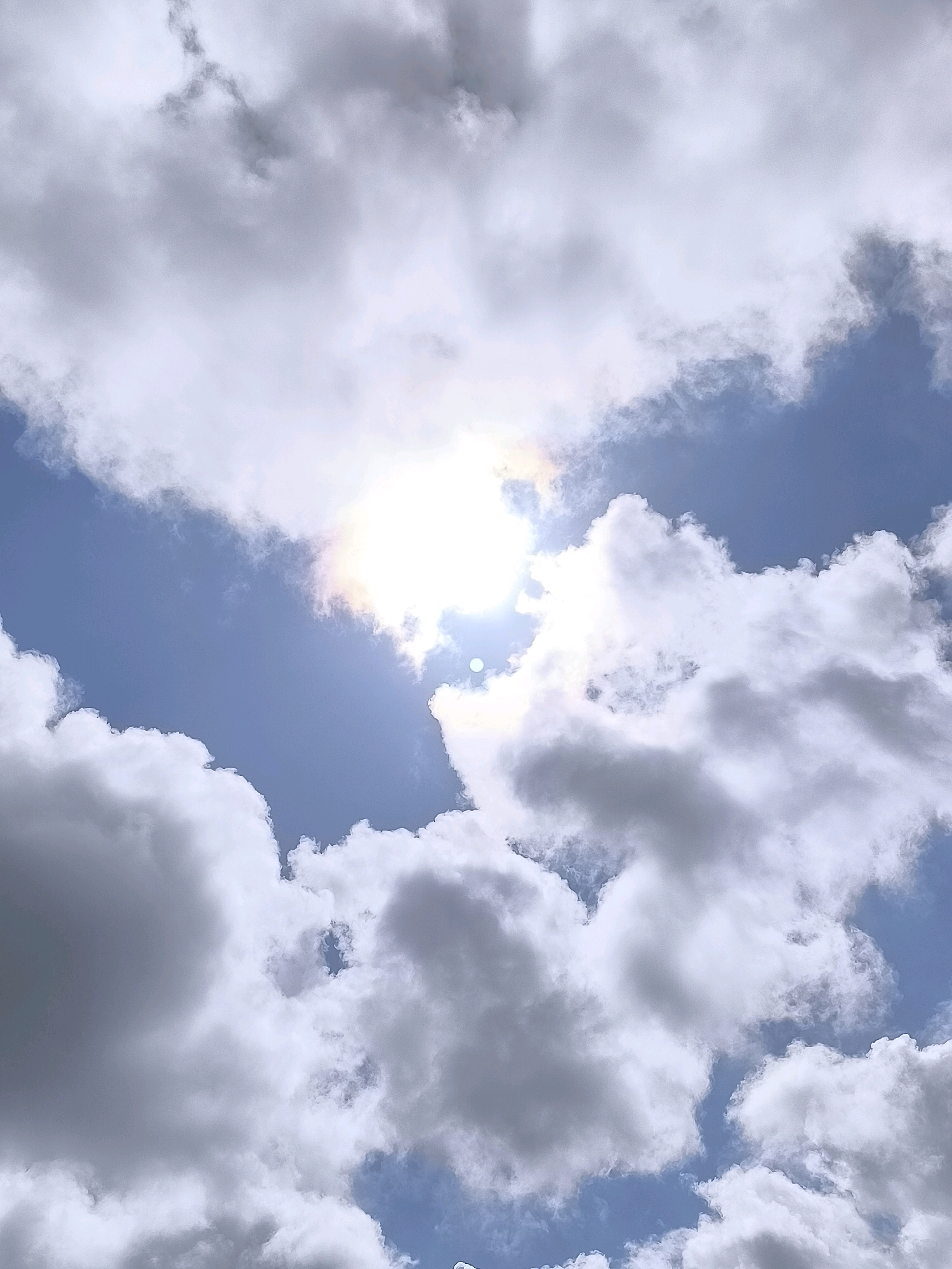This image displays a close-up, low-angle view of a partly cloudy sky. The sun is visible, partially obscured by clouds, creating a bright, hazy effect with a subtle rainbow aura. The clouds themselves are a mix of bright white and darker gray, suggesting varied thickness and density. The background sky is a clear, light blue. The scene is not specific to any particular location within the city of Laistai, Lithuania, as it is a generic sky composition without any recognizable landmarks or urban elements.