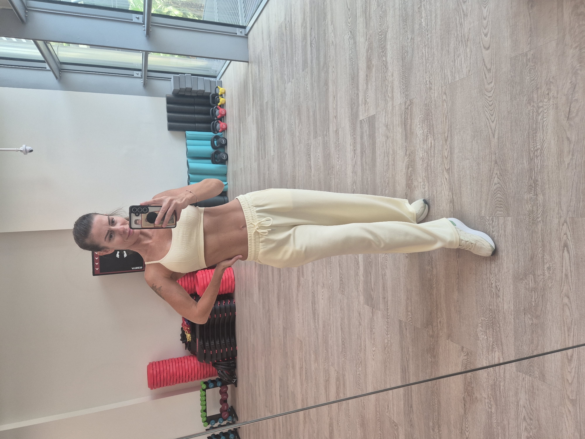 The image shows a woman in a gym, standing in front of a mirror and taking a selfie. She is wearing a light-colored ribbed sports bra and matching sweatpants, along with white sneakers. The gym has light-colored wooden floors and white walls. Exercise equipment, including yoga mats, dumbbells, kettlebells, and foam rollers, can be seen in the background along the walls. A large window with a view of trees outside is visible in the upper part of the frame. The location is a gym in an unknown city and location.
