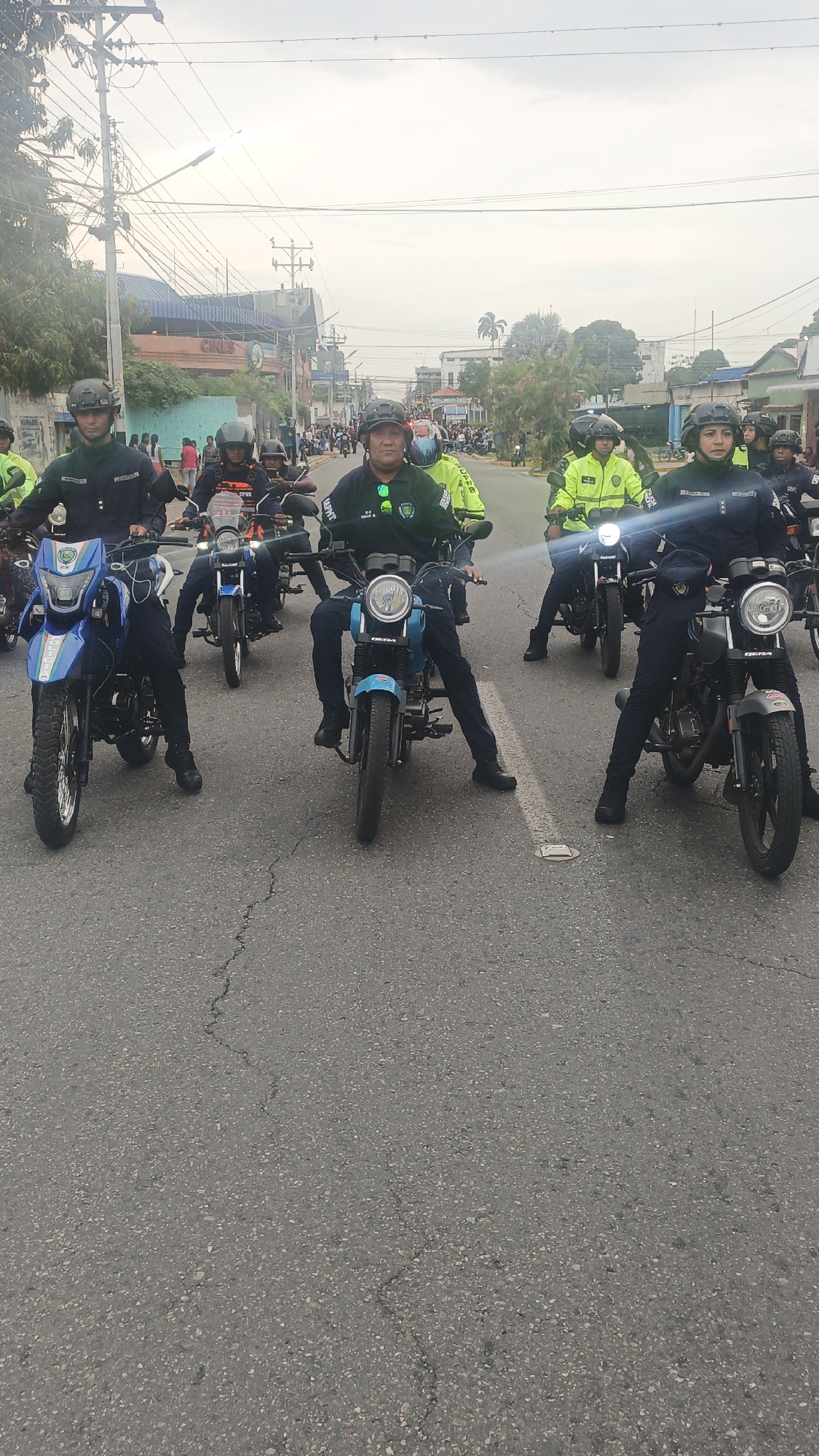 The image shows a group of uniformed police officers on motorcycles lined up on a paved road, seemingly waiting or participating in a procession. The scene is taking place in Tinaquillo, Venezuela, which can be deduced from visible text on the officers' uniforms. The officers are wearing helmets and dark green or blue uniforms. Some of them are wearing yellow vests. The motorcycles are various models and colors, including black, blue, and silver. In the background, there are buildings, overhead power lines, and a crowd of people gathered on the sidewalks. The sky is overcast.