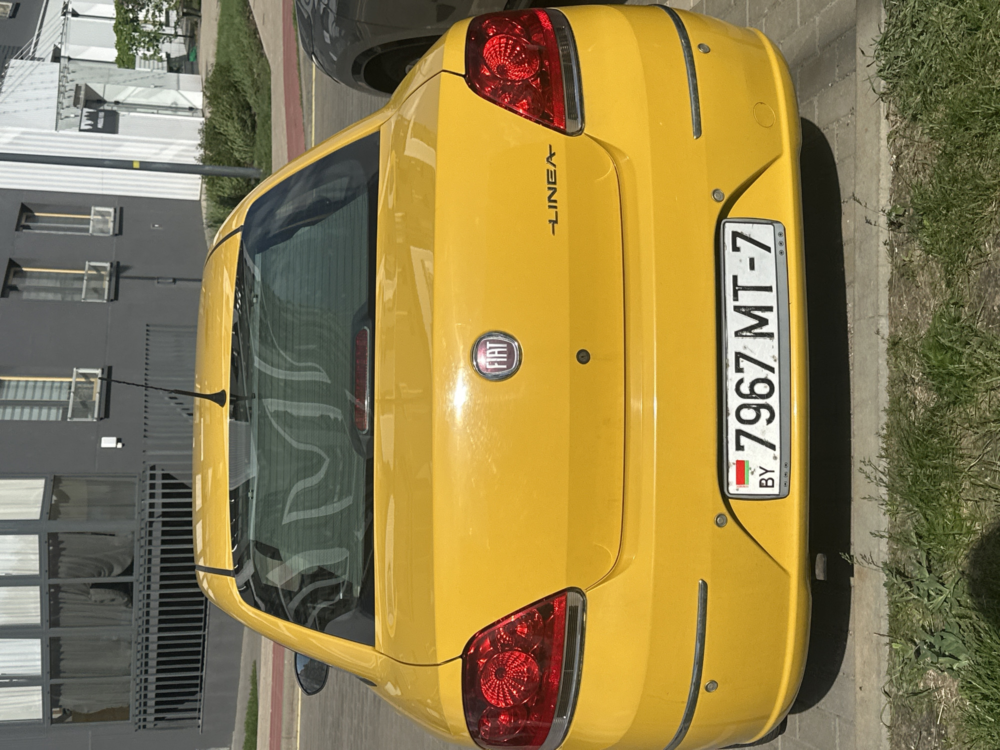 A yellow Fiat Linea is parked on the side of a street in Бараўлянскі сельскі Савет, Belarus. The car's rear is visible, showcasing its license plate, "7967 MT-7 BY". The building and fence are on the left, and grass and a road are on the right.