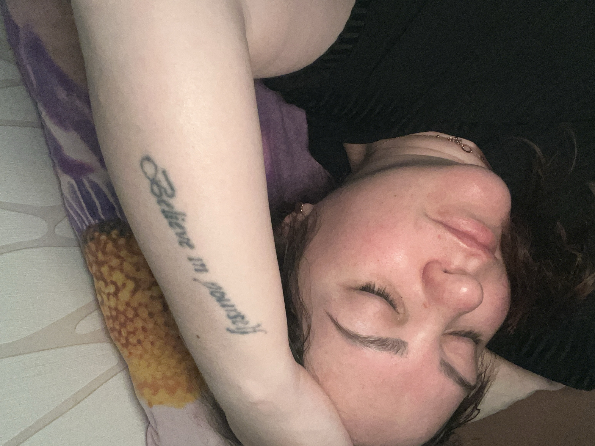A woman is lying on her side, eyes closed, seemingly asleep. Her left arm is bent and positioned near her head; a tattoo reading "Believe in yourself" is visible on her forearm. She is wearing a black top. Her head rests on a pillow with a floral design. A necklace is visible on her neck. The location of the image is unknown, so I cannot confirm it is Perm, Russia.