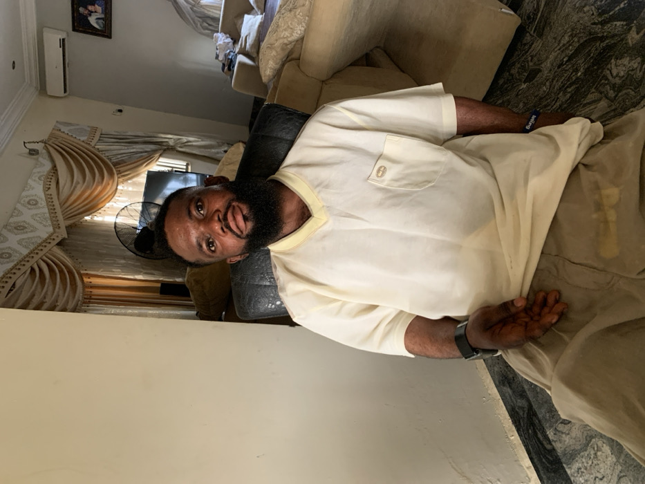 The scene depicts an adult male seated in an indoor living room setting in Eika-Adagu New Layout, Nigeria. The subject, a dark-complexioned male with a full beard and short dark hair, is wearing a light cream V-neck short-sleeved shirt with a left chest pocket featuring a small brown emblem, and light brown trousers. A black smartwatch is on his left wrist, and a blue wristband is on his right. He is seated in a dark, possibly leather, armchair, looking directly at the camera with a neutral expression.
The background shows light-colored walls and a floor tiled with a grey and white marble-like pattern. A light-colored sofa is partially visible to the right. Behind the subject, a window is covered by elaborate light gold/beige curtains with ornate valances. A black standing fan is positioned to the left of the curtains, in front of a partially visible flat-screen television. A wall-mounted air conditioning unit and a framed picture are visible on the wall to the right of the curtains. A light-colored wall partition is present on the left side of the image. The subject is stationary, posing for the photograph.