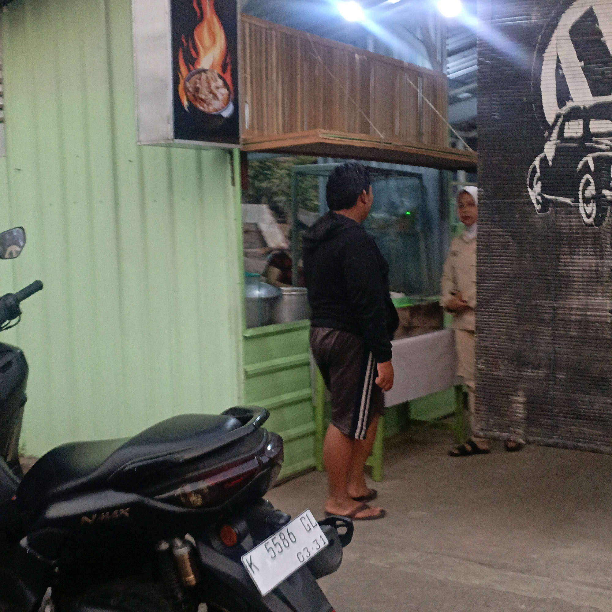 The image depicts an outdoor scene in Salatiga, Indonesia, likely in front of a food stall or small restaurant. In the foreground, a black motorcycle is parked, with its rear visible. The license plate reads "K 5586 GL 03-31". To the left, a light green corrugated metal wall forms the backdrop.
In the middle ground, a man wearing a black hooded sweatshirt and dark shorts stands facing away from the camera, looking towards the right. To his right, a woman wearing a light-colored headscarf and a matching outfit is partially visible. She appears to be looking towards the man.
Behind the people, there's a structure with wooden slatted walls, possibly an elevated seating area or part of the food stall's construction. A sign with a flame motif and an image of food is mounted on the left wall. To the far right, a large black graphic featuring a vintage car is affixed to a textured surface, likely a wall. The environment appears to be dimly lit, with some overhead lighting visible. The ground is paved.