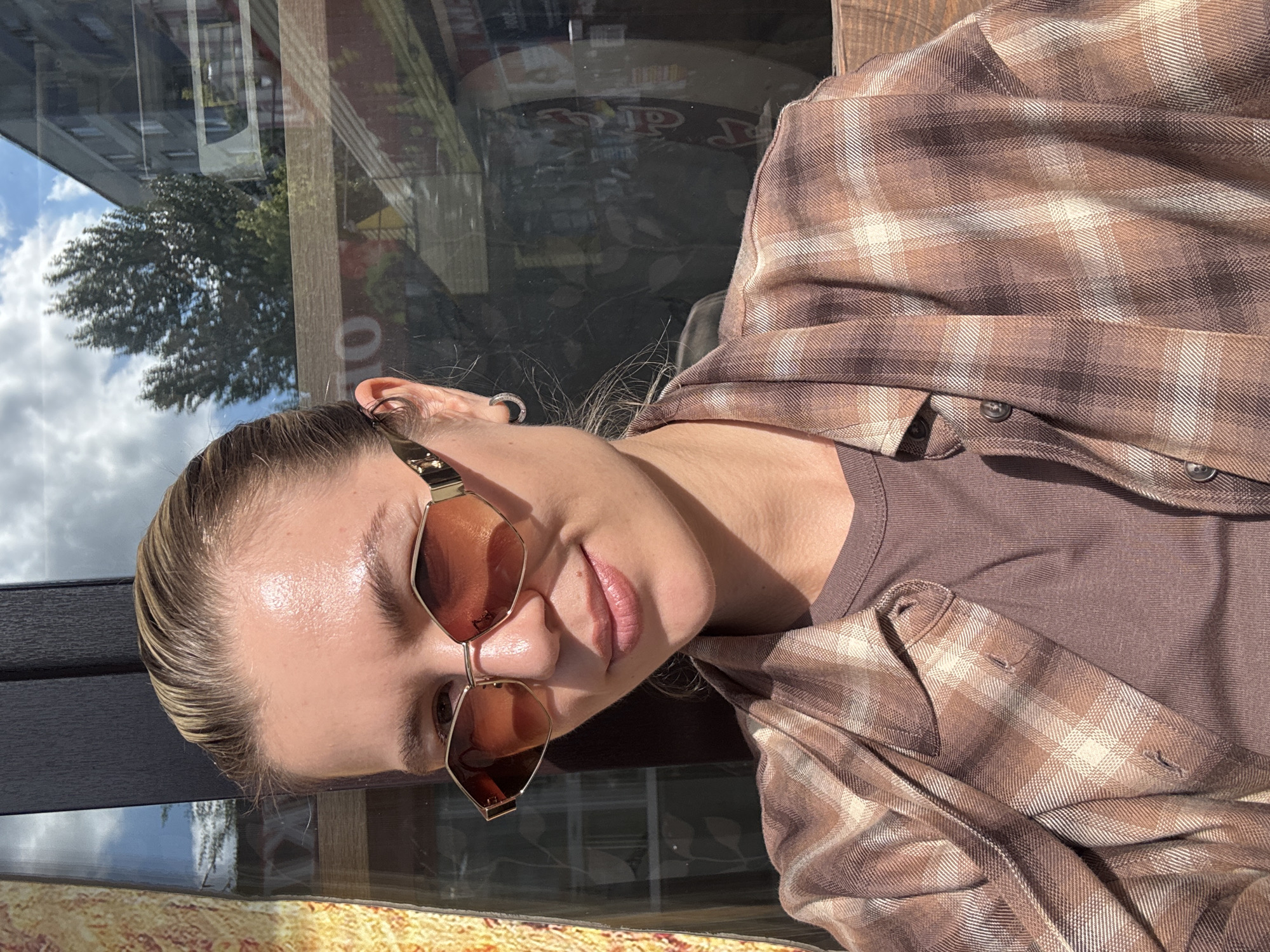 A woman with blonde hair pulled back and wearing sunglasses is taking a selfie outdoors. She is wearing a brown plaid shirt over a brown t-shirt. The background shows a reflection in a window of trees, a building, and a sign with "TO" visible. The lighting suggests it is daytime.