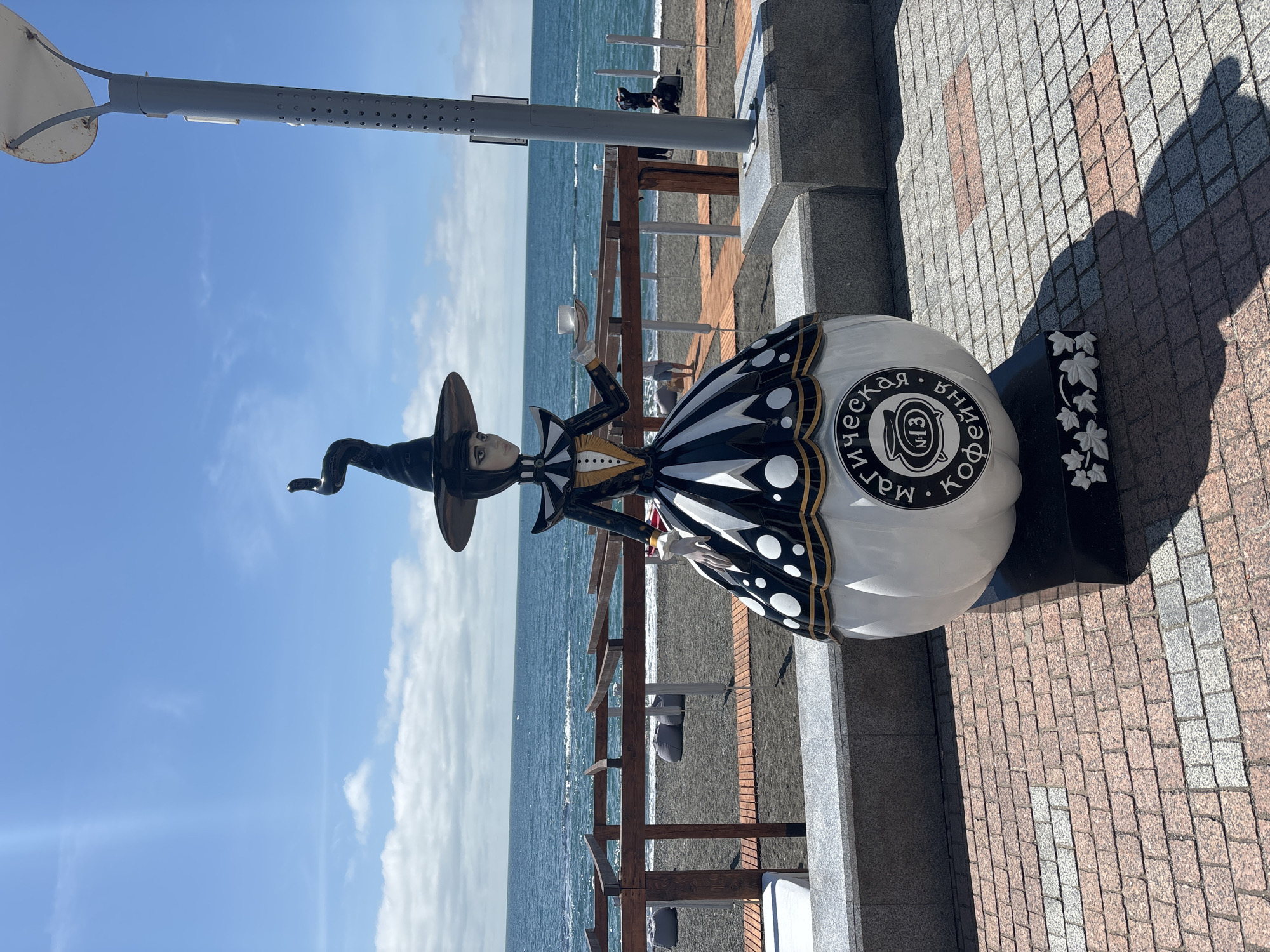The photograph captures a whimsical sculpture of a witch positioned along a paved embankment in the city of Sochi, Russia. The sculpture is strikingly detailed, featuring a woman in a black witch's hat and a black-and-white patterned dress. The base of the sculpture is shaped like a white orb, adorned with black lettering and an emblem containing the number 13 and a coffee pot image. A black base with floral carvings sits beside it.
The embankment runs parallel to a dark-sand beach and a serene body of water, presumably the Black Sea. A wooden railing separates the embankment from the beach. In the background, figures of people can be discerned on the beach, though they are too distant to identify.
A tall, grey lamppost stands adjacent to the sculpture. The sky is a bright blue, dotted with sparse clouds. The overall scene suggests a sunny, pleasant day at the seaside.