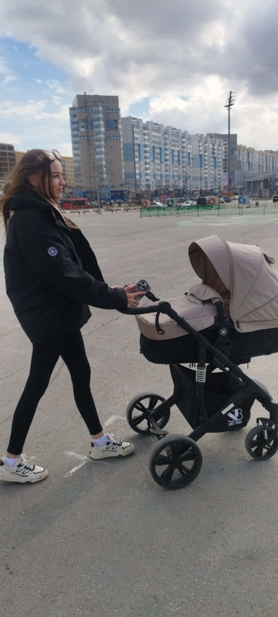 The photograph depicts a young woman walking with a baby stroller in what appears to be a public square or parking lot. The location is identified as Yakutsk, Russia based on the architecture and urban setting visible in the background. The woman is wearing a black puffy jacket, black leggings, and white sneakers with white socks. She has long brown hair and is wearing sunglasses on her head. She is holding a smartphone in her right hand, which is attached to the stroller's handle. The stroller is a dark-colored "Sweet Baby" brand model, with a beige canopy and storage compartment. The surrounding environment includes a large paved area, with tall apartment buildings in the background, characteristic of urban residential areas. There are various urban features visible, like light poles, parking areas, and abstract circular green and white painted spots. The sky is overcast.