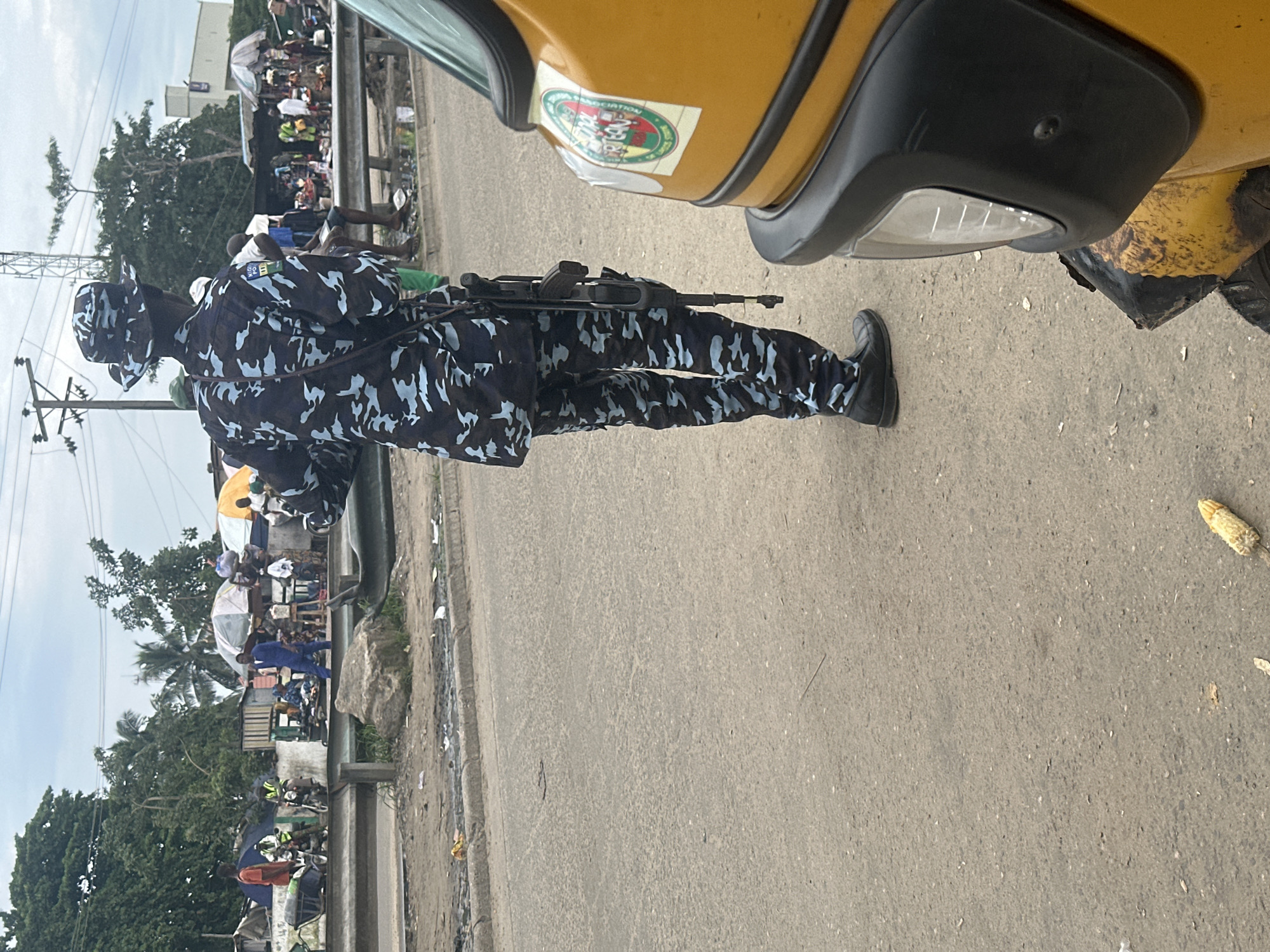 A Nigerian police officer in camouflage uniform with a blue and light blue pattern stands facing away from the camera in the foreground. The officer is wearing a camouflage hat and is equipped with a rifle slung over their right shoulder. To the right of the officer, a portion of a yellow motorized tricycle is visible, featuring a logo on its side. The scene appears to be a roadside market in Badagry, Nigeria, with a busy street and numerous stalls under makeshift tents. In the background, people are walking and interacting, with various vehicles, including motorcycles, present. The environment is dusty and appears to be an outdoor, open-air market.