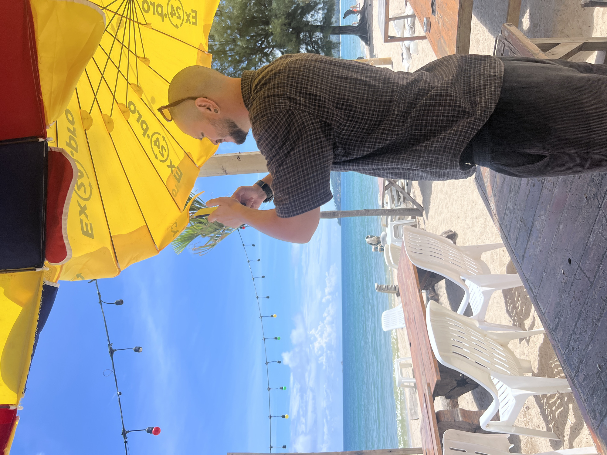 A bald man wearing a plaid shirt and dark pants is standing on a beach in Choeng Thale, Thailand, holding a phone up to take a picture of some greenery. He is standing under a large yellow umbrella that has "EX(s)" printed on it in a repeating pattern. The beach has white plastic chairs and wooden tables set up, with the ocean in the background. The sky is blue with scattered white clouds. String lights are visible overhead.