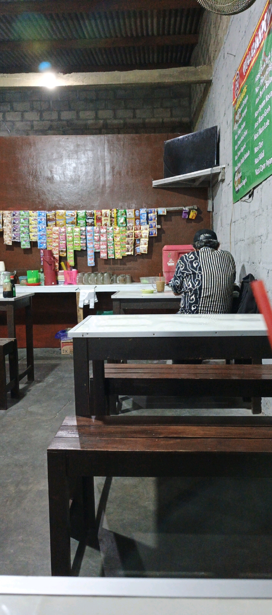 The image depicts the interior of a casual dining establishment, likely a warung or small restaurant, in Banyuraden, Indonesia. The space is dimly lit, with a corrugated metal ceiling and brick walls. In the foreground, dark wooden tables and benches are visible, suggesting a communal dining setup. A person, viewed from behind, is seated at one of the tables, wearing a black and white striped shirt and a black cap. The person appears to be engaged with something on the table.
The middle ground reveals a counter area with various items displayed. A prominent feature is a long row of individually packaged snacks and instant drink mixes hanging on the wall above the counter. On the counter itself, there are glasses, a red thermos, a pink basket, and some condiment bottles. A red cooler is also visible behind the counter.
In the background, a flat-screen television is mounted on the wall. The overall atmosphere suggests a simple, local eatery. The presence of a menu board on the far right wall, with handwritten text in Indonesian, further confirms the establishment's nature. The environment is clean but utilitarian, indicative of a no-frills dining experience catering to the local community.