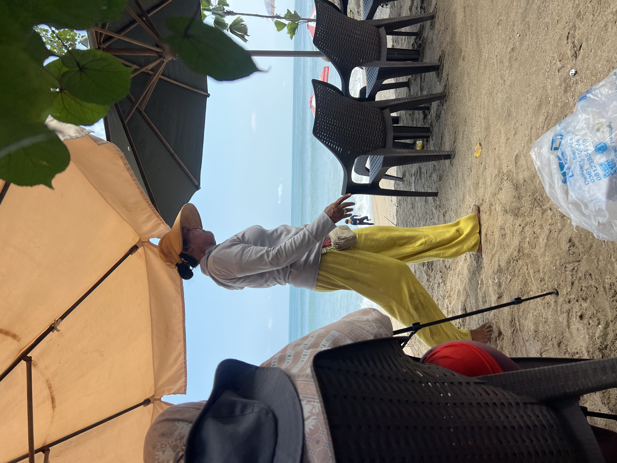 On a sandy beach in Kuta, Bali, Indonesia, a woman wearing a light-colored long-sleeved shirt and bright yellow pants walks along the shore. She has a yellow hat on and a small tan pouch hanging from her waist. In the background, the turquoise ocean meets a clear blue sky. Several dark plastic chairs are set up on the sand, and two beach umbrellas, one orange and one dark green, provide shade. A person in a dark outfit is visible near the water's edge. In the foreground, a chair is partially visible on the left, and a white plastic bag lies on the sand. The scene appears to be a typical beach setting with a mix of leisure and service elements.
