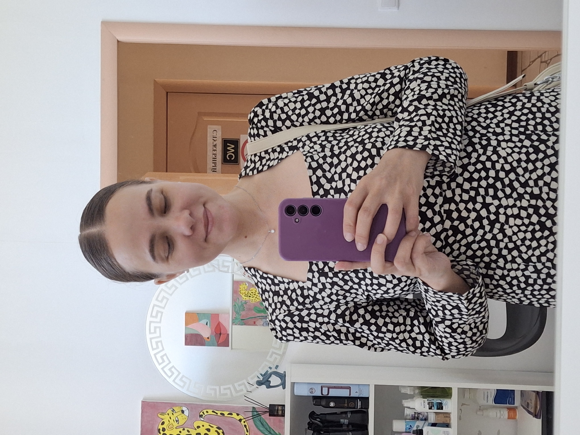 A woman with dark hair is taking a selfie in a mirror. She is wearing a black and white patterned dress and holding a purple smartphone. The background includes a door with Russian text "СЛУЖЕБНИЙ МС," a shelf with various items, and artwork featuring a leopard. This image was taken in the city of Chaikovsky, Russia.