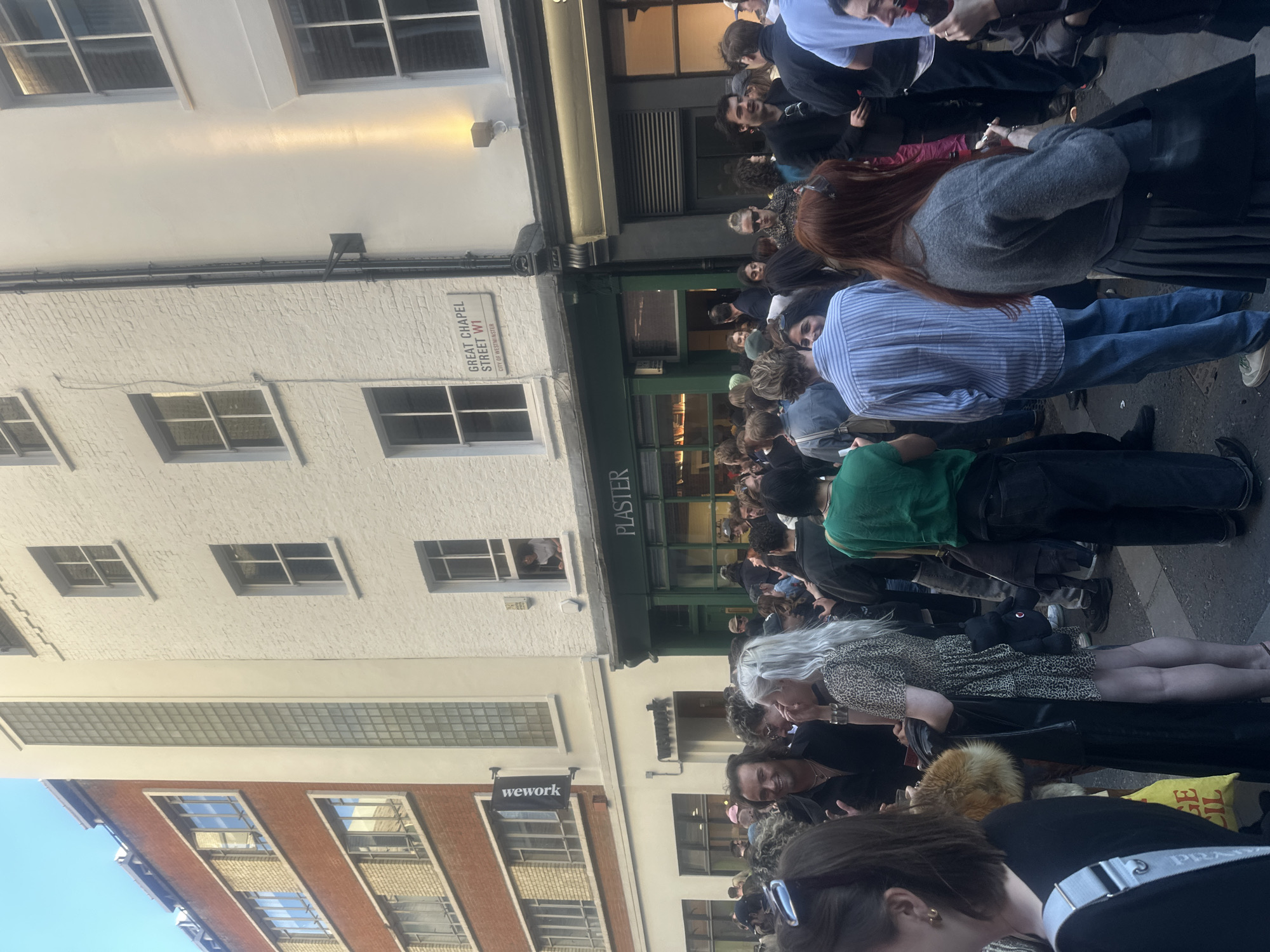 A daytime street scene in London, United Kingdom, displays a dense crowd of people congregated on Great Chapel Street, W1. The street features buildings with architectural signage, including "PLASTER" on a green facade and a "wework" sign. The crowd is composed of individuals in varied attire; some are standing and interacting, while others appear to be looking in the same direction. The image conveys a sense of urban activity and congestion in this particular area.