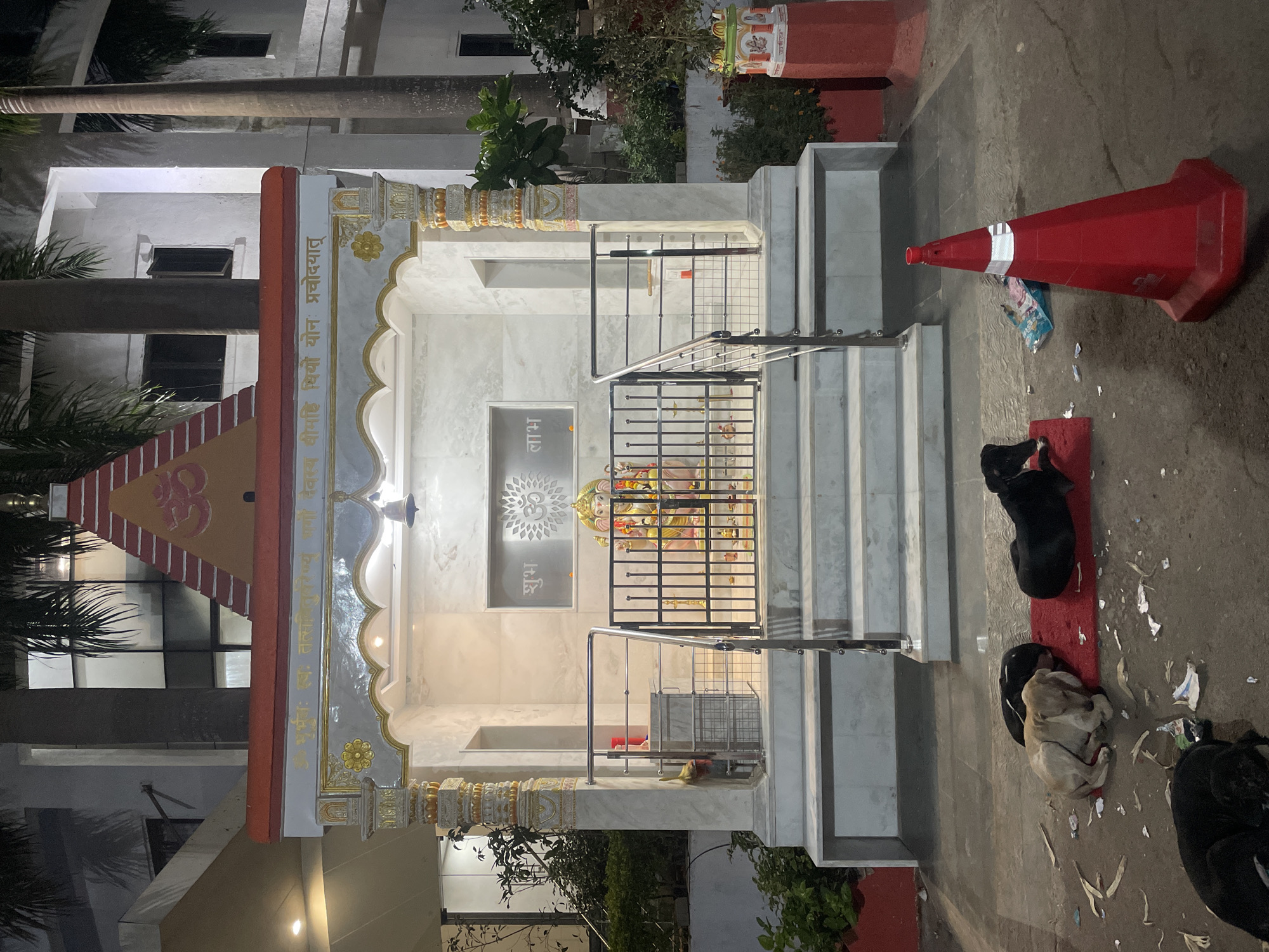 The image depicts a small Hindu temple at night. The temple has a light-colored marble or stone structure with decorative columns and a roof adorned with Hindu symbols and inscriptions. Inside, a deity is visible behind a metal gate. Outside the temple, on the ground, are several dogs, some lying on a red mat. A red traffic cone is also visible near the dogs. The location is likely Mumbai, India, based on the style of the temple and the presence of street dogs, a common sight in Indian cities.