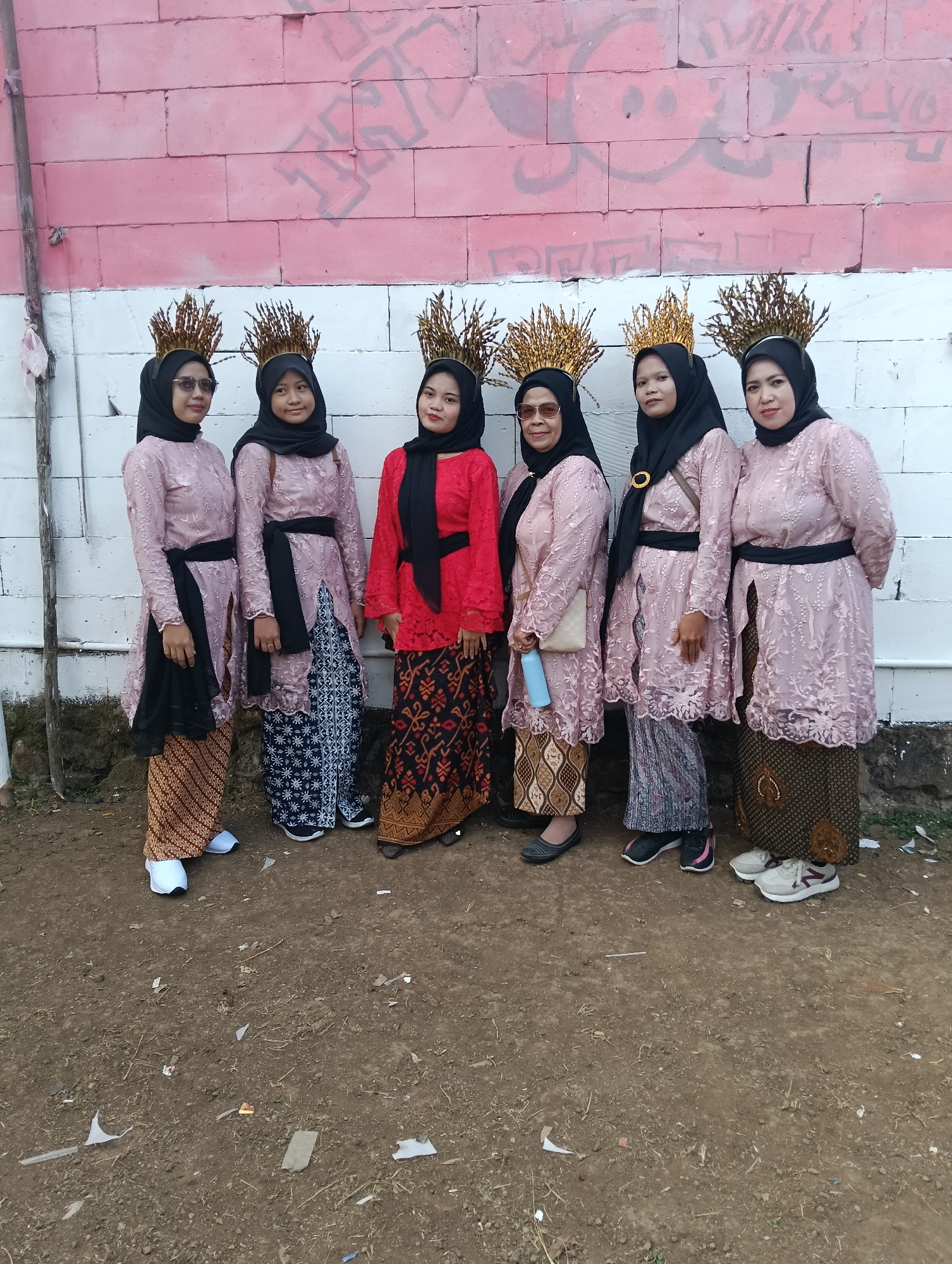 Six women, dressed in traditional Indonesian attire, stand side-by-side in front of a pink and white brick wall covered in graffiti. Each woman wears a golden, crown-like headpiece made of what appears to be dried rice stalks. Their clothing consists of long, pink, lace-like tunics with black sashes tied around their waists, paired with patterned skirts or trousers. The woman second from the left is wearing a red top and a patterned skirt. The ground in front of them is dirt, scattered with debris. The background wall has a rough, unfinished texture. This scene likely takes place in Tasikmalaya, Indonesia, given the traditional clothing and cultural context.