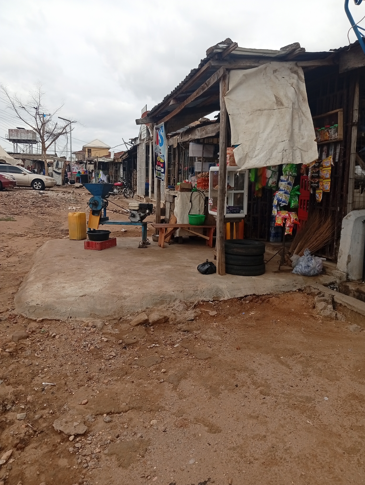 This media file captures a bustling roadside market scene in Zango-ndaji, Nigeria, under an overcast sky, suggesting daytime. The foreground features an uneven, reddish-brown dirt ground with scattered pebbles, leading up to a raised, weathered concrete platform.
On this platform, a prominent blue, engine-powered grinding machine with a yellow hopper is set up, implying a service for processing grains or other foodstuffs. A black basin and a red plastic crate are placed beneath its chute, while a yellow jerrycan rests nearby. Adjacent to the machine, a small wooden bench, three stacked black tires, and a black plastic bag are visible. Further along the platform, a bundle of brooms and a clear plastic bag containing items lean against the main structure.
The main subject is a rustic market stall or shop, constructed with wooden poles supporting a corrugated metal roof. A large, off-white tarp hangs from the front, partially obscuring the interior. Displayed within and around the stall are various packaged snacks like crisps and biscuits, hanging from hooks or arranged on shelves. A white display cabinet or cooler is also visible.
In the middle distance to the left, a beige sedan and a red car are parked on the dirt road. Behind them, a bare-branched tree stands next to a tall structure topped with a water tank. Utility poles and wires crisscross the sky. The background reveals a row of similar makeshift market stalls or buildings extending down the road, with indistinct figures of people moving about. A red motorcycle is also visible further back.
A blue banner-like sign hanging from a distant stall shows the letters "TS," likely part of a brand or shop name. The overall scene depicts a typical commercial area, likely offering various goods and services to the local community.