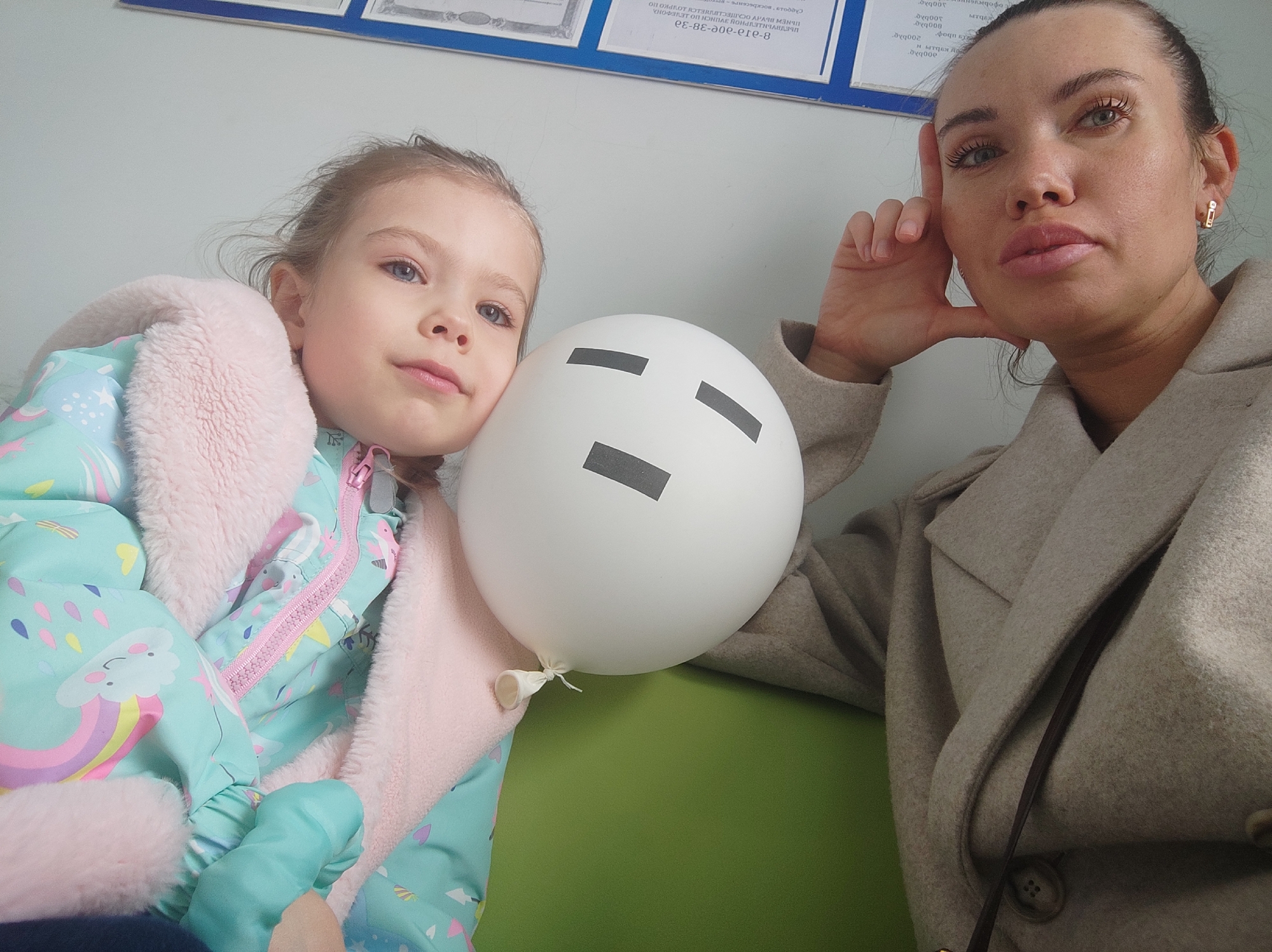 A woman and a young girl pose for a photo in what appears to be an indoor waiting area. The woman is on the right, wearing a light beige coat, her dark hair pulled back, and she is resting her head on her hand while looking directly at the camera. The young girl is on the left, wearing a blue winter coat with a pink fur trim, and she is leaning against a white balloon with a minimalist face drawn on it. The background includes a document frame on the wall, with partially legible text, which seems to indicate that the location may be in the city of Votkinsk, Russia. The scene is lit with what appears to be artificial lighting.