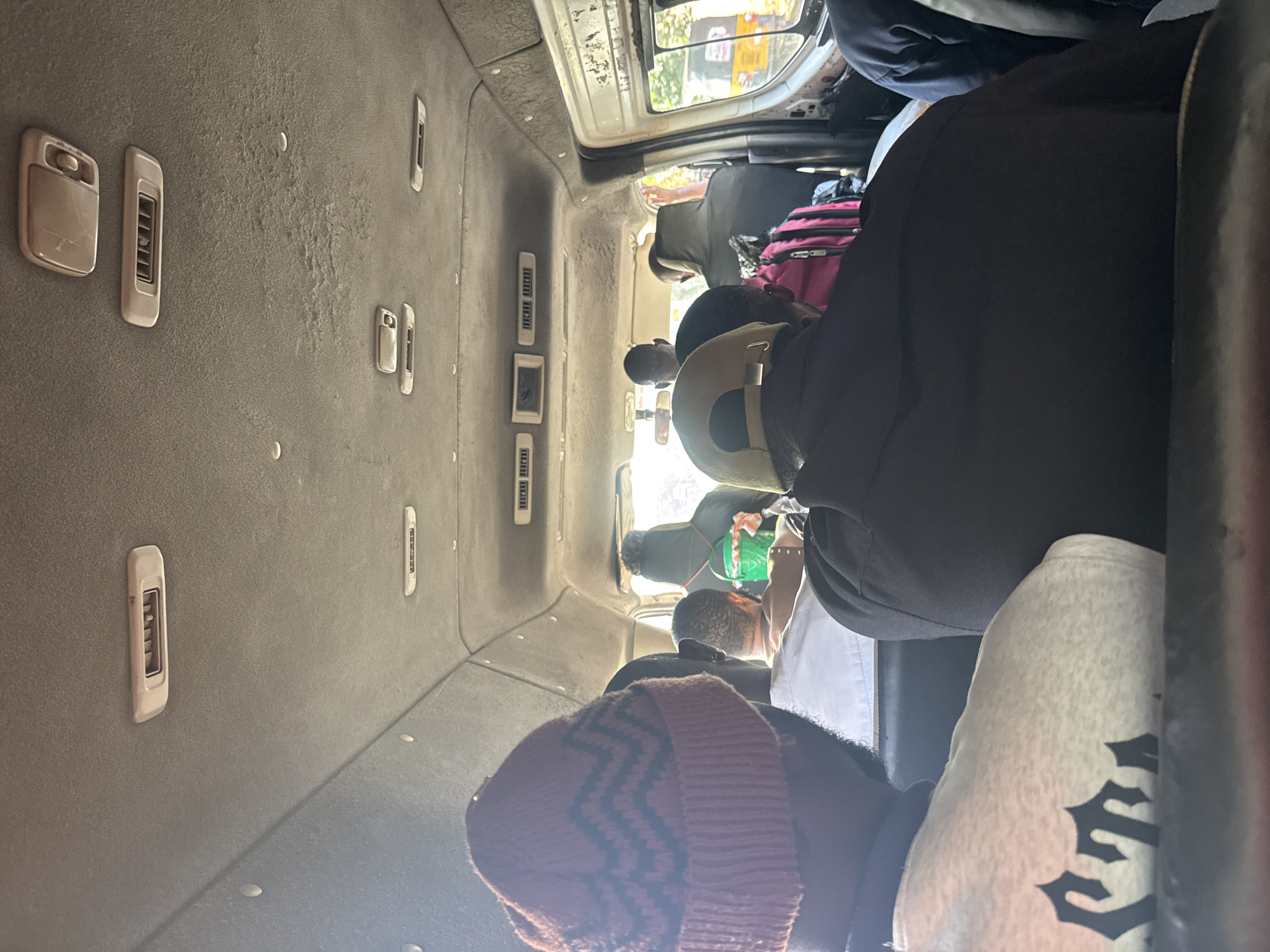The image depicts the interior of a crowded passenger van, likely a public transport vehicle, somewhere in the city of Orifite, Nigeria. Several individuals are visible seated close together, with a range of clothing and headwear apparent including a rust-colored beanie. The van's interior features air vents along the ceiling. Luggage and other personal items are stored above the seats. The lighting suggests a bright, possibly sunny, day.
