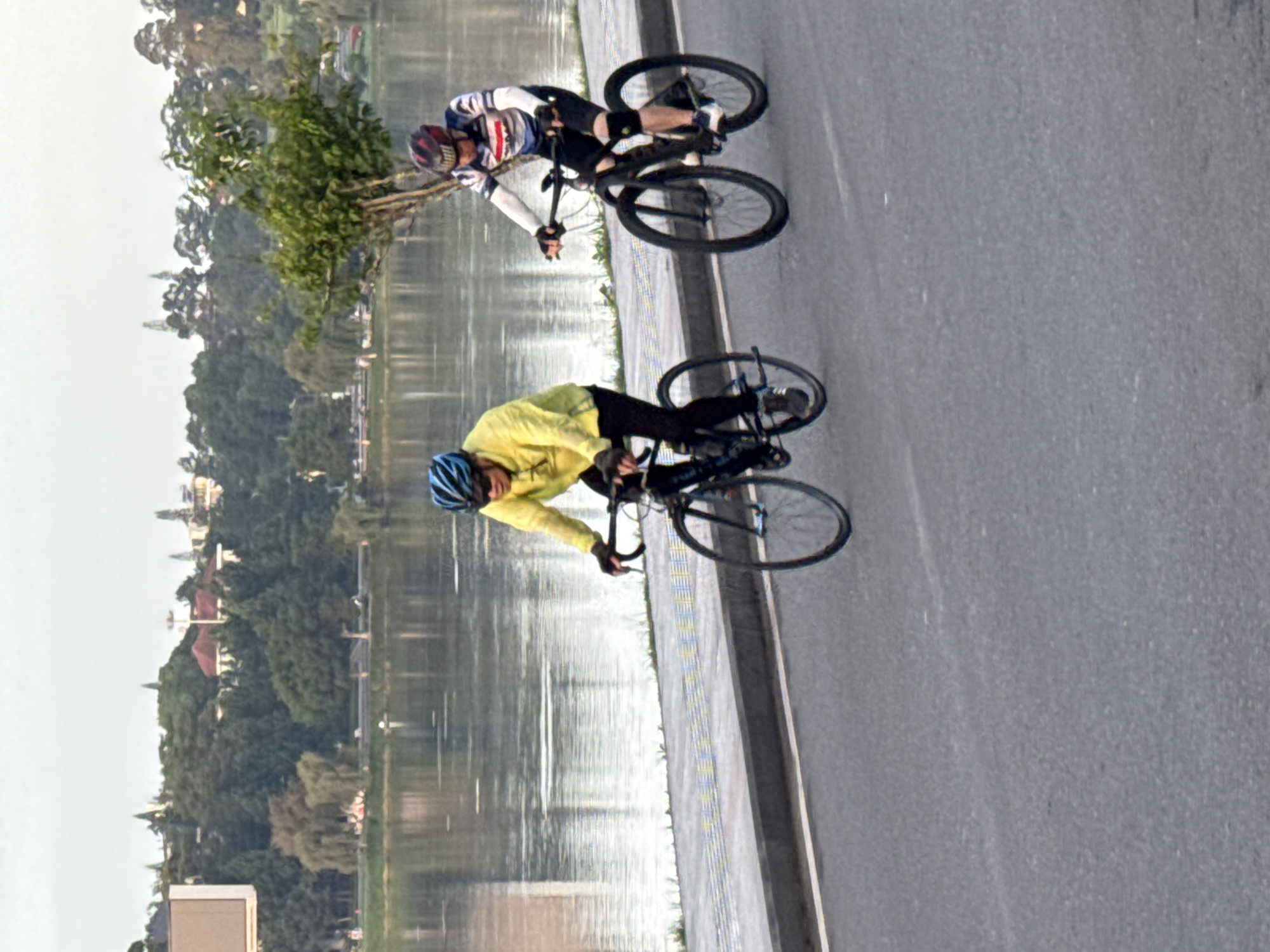 Two cyclists are riding on a paved road next to Xuan Huong Lake in Da Lat, Vietnam. The cyclist in the foreground is wearing a yellow long-sleeved shirt and black pants, with a blue helmet. The cyclist behind is wearing a blue and white striped shirt with black shorts and a red helmet. Both cyclists are wearing helmets and appear to be engaged in recreational cycling. The lake is calm and reflects the overcast sky. In the background, lush green trees and some buildings are visible on the far bank of the lake, with a billboard on the left. The road surface is asphalt, and there is a sidewalk or pedestrian path adjacent to the road with a decorative border. The overall impression is a serene outdoor scene in a city park setting.