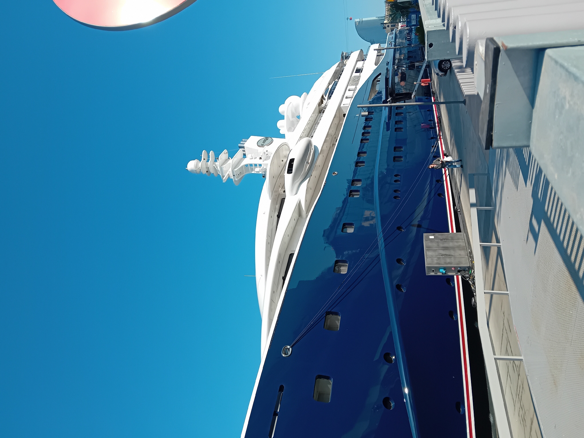 The image displays a large, luxurious yacht or cruise ship docked at a port under a clear, bright blue sky. The ship's hull is a deep, glossy blue, accented by a distinct red and white stripe running along its waterline. Its expansive superstructure is white, featuring multiple decks, numerous windows, and sophisticated equipment such as radars, antennae, and life rafts or tenders on its upper levels.
One person is visible standing on the concrete dock adjacent to the ship, dressed in a dark jacket and light trousers. Further down the pier, a white SUV-style car is parked. The dock itself is equipped with grey railings, light poles, and several utility boxes, including a black one with visible blue and red connectors. The presence of strong, well-defined shadows cast by the railings and the ship indicates bright, sunny conditions, suggesting it is mid-morning or afternoon. The sky is entirely cloudless.
In the upper left portion of the image, a partial, light-colored, and reflective circular object is visible, possibly part of nearby infrastructure or another vessel. On a white circular housing located on the ship's superstructure, the text "AL MAHARA" is discernible. The overall scene captures a serene and sunny day at a port in Barcelona, Spain.