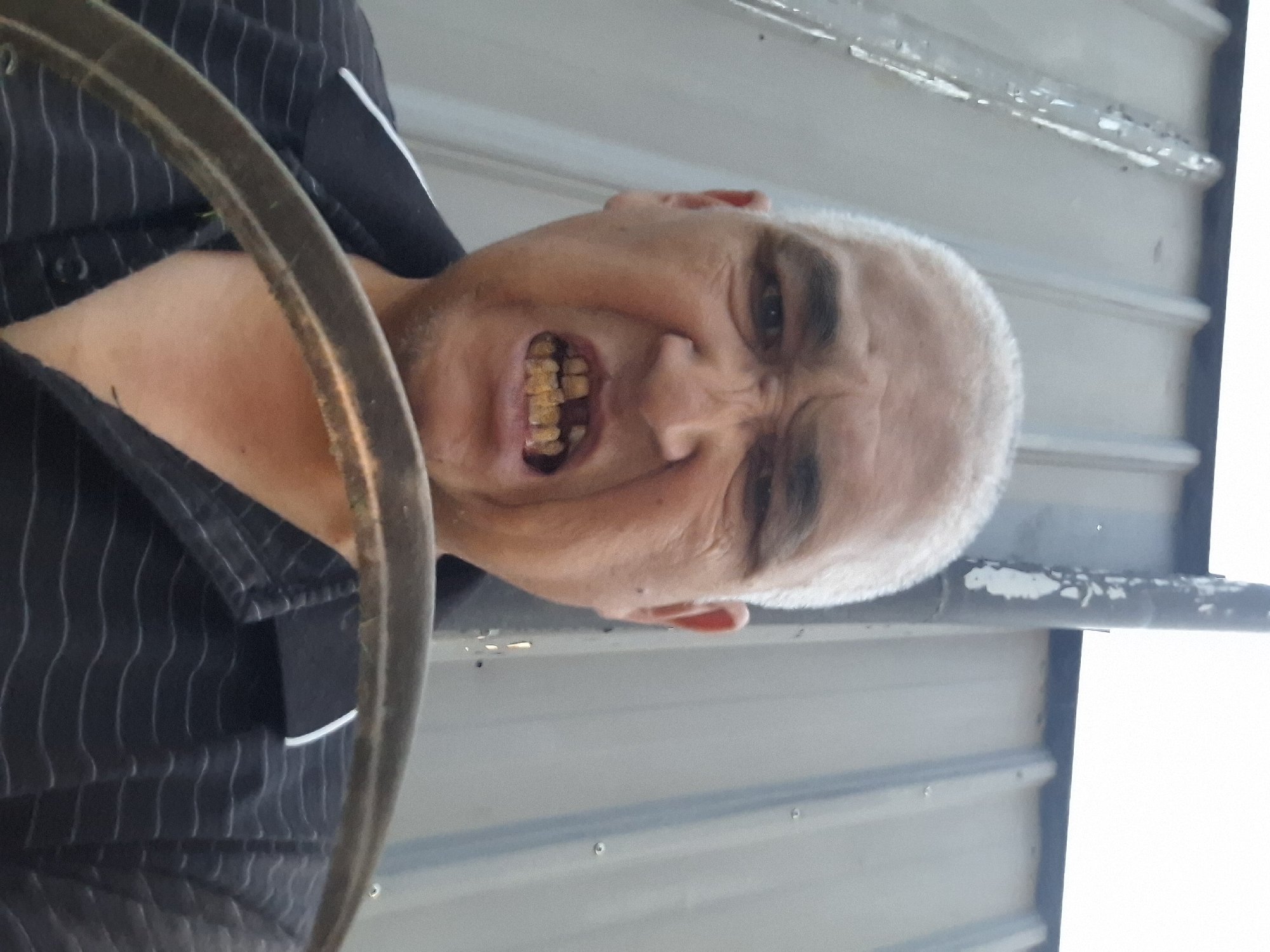 A low-angle, head-and-shoulders selfie taken outdoors. The subject, an adult male with light skin and a shaved head, is looking directly at the camera with an aggressive facial expression. His mouth is open, revealing discolored and broken teeth. He is wearing a dark collared shirt with thin white pinstripes. In the foreground, a circular metal object, possibly a steering wheel or part of a tool, obscures the lower portion of his chest. The background consists of a corrugated metal wall, appearing to be part of a building or fence, with a vertical pipe on the right. The lighting suggests it is daytime, possibly overcast. There is no discernible information to place this image in Almaty, Kazakhstan.