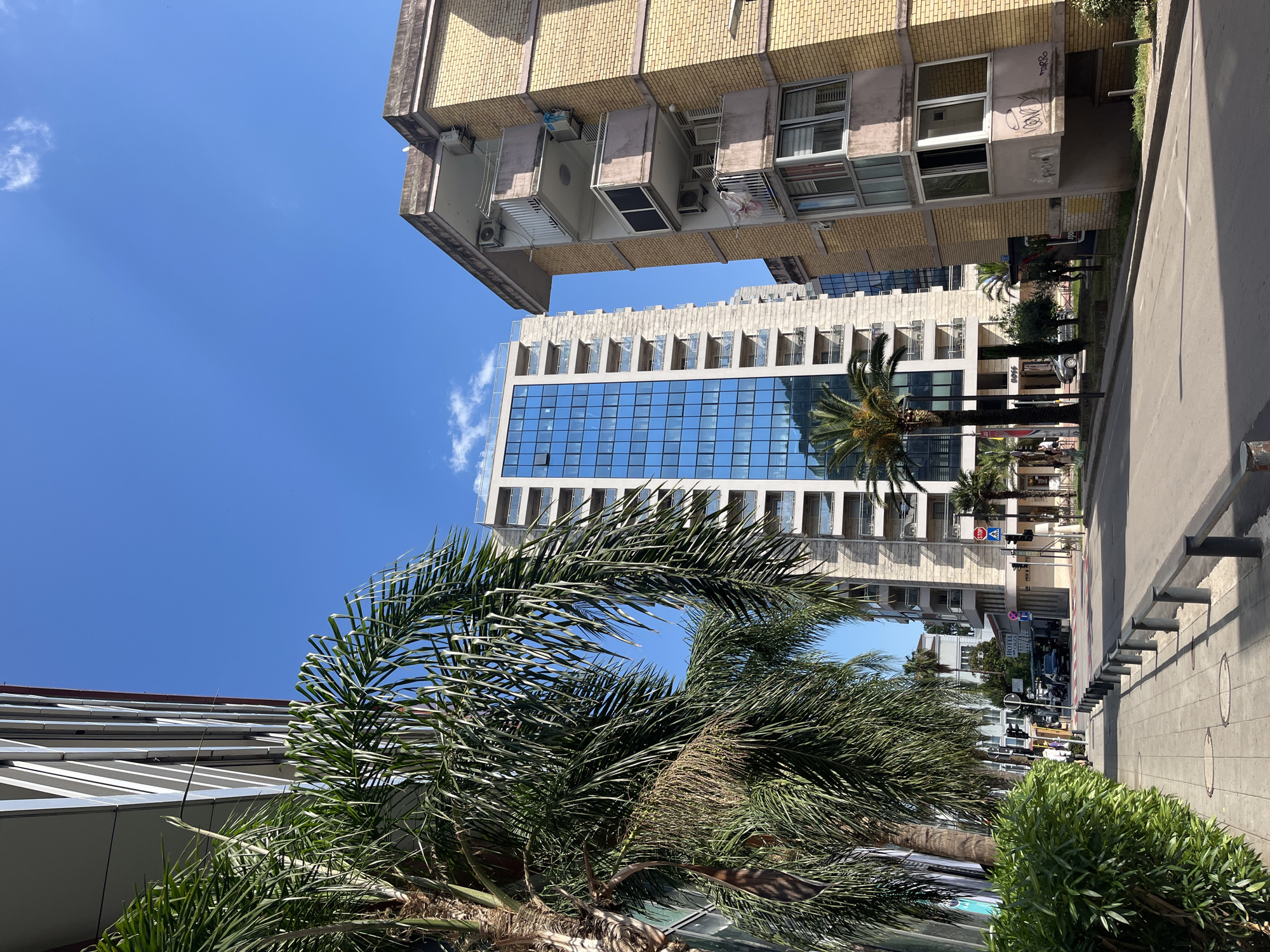 A low-angle, eye-level shot captures a street scene in Budva, Montenegro, on a clear, sunny day. Palm trees with lush green fronds flank the left side of the image, partially obscuring a modern building with a sleek facade composed of reflective blue glass and white horizontal accents. To the right, a contrasting building with yellowish-brown brickwork stands prominently, featuring multiple balconies with light-colored railings and some with enclosed windows.
The street itself is paved with grey cobblestones in the foreground, transitioning to asphalt further down. A metal railing separates the sidewalk from the street. Several vehicles are visible parked along the street and on the road, including cars and a bus. A traffic light with a red signal is positioned on the right side of the intersection. Signs such as "STOP" and "HOTEL" are discernable on buildings.
A few individuals are present on the sidewalk, one walking and others appearing to be near shop entrances. The sky is a vibrant blue with a few wispy white clouds. The overall impression is of a lively urban environment with a mix of modern and older architectural styles, characteristic of a coastal city.