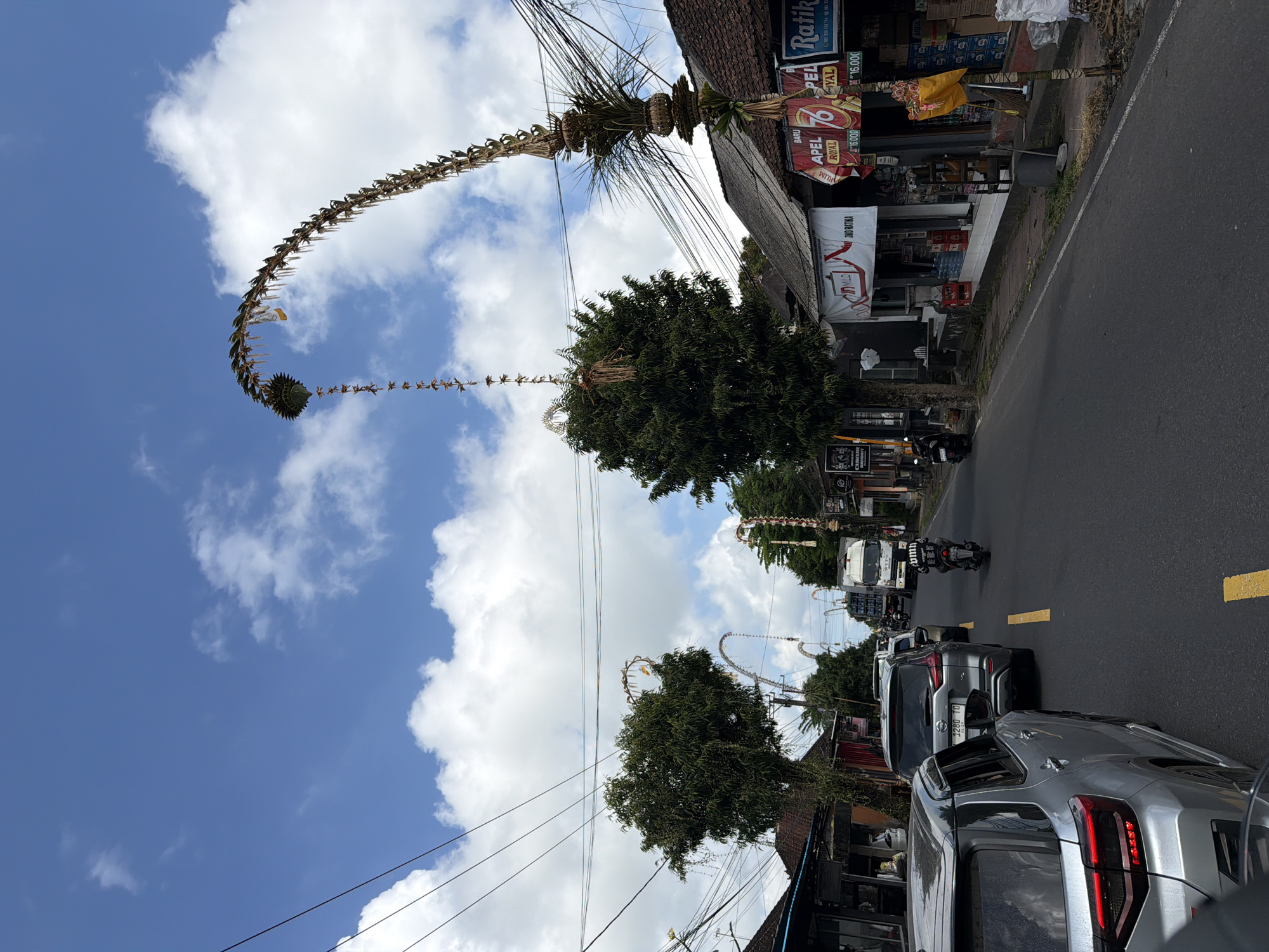 The image depicts a street scene in Tampaksiring, Indonesia, with a narrow road bordered by shops and lush green trees under a bright blue sky dotted with white clouds. Several vehicles, including cars and a motorcycle, are present on the road, suggesting moderate traffic. The shops lining the street display various advertisements and products, with some featuring festive decorations, possibly related to a local celebration. Large, elaborately crafted bamboo and palm leaf structures, known as "penjor," arch over the road, adding to the culturally rich atmosphere. These decorations are characteristic of Balinese traditions. The overall environment suggests a bustling, yet culturally vibrant, urban or semi-urban setting.