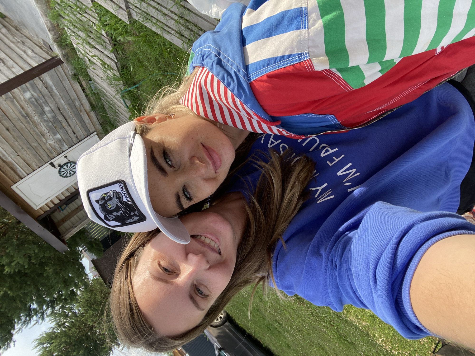 Two women are taking a selfie outdoors. The woman on the left has long brown hair and is wearing a blue sweatshirt. The woman on the right has blonde hair, is wearing a white baseball cap with a black panther logo, and a colorful striped jacket. They are smiling and appear to be in a rural or backyard setting. In the background, there is a wooden structure, some trees, and a grassy area. This scene is consistent with the exterior of a house or dacha in a dacha settlement like the one in Garevaya, Russia.