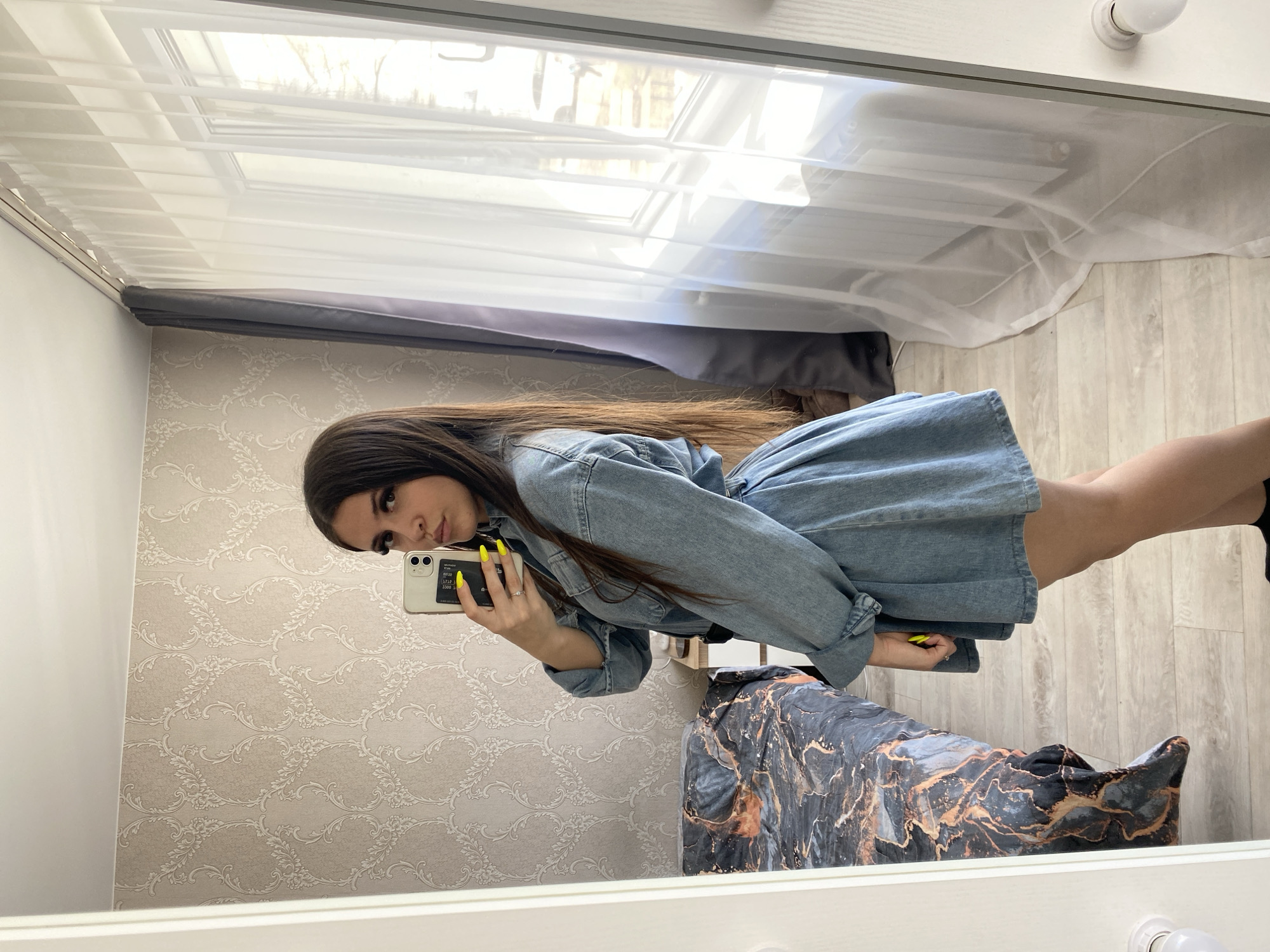 A woman with long, dark brown hair is taking a selfie in a mirror. She is wearing a light blue denim dress and has brightly colored yellow nails. In her left hand, she is holding a white smartphone, presumably taking the picture. The background, reflected in the mirror, features a wall with a beige and white floral pattern. There's also a window with white blinds and sheer white curtains, and a grey valance above the window. In the bottom right of the reflection, there is a patterned garment bag. The image was captured in what is likely a private indoor space. The city of Chaikovsky, Russia is not directly indicated in the image.