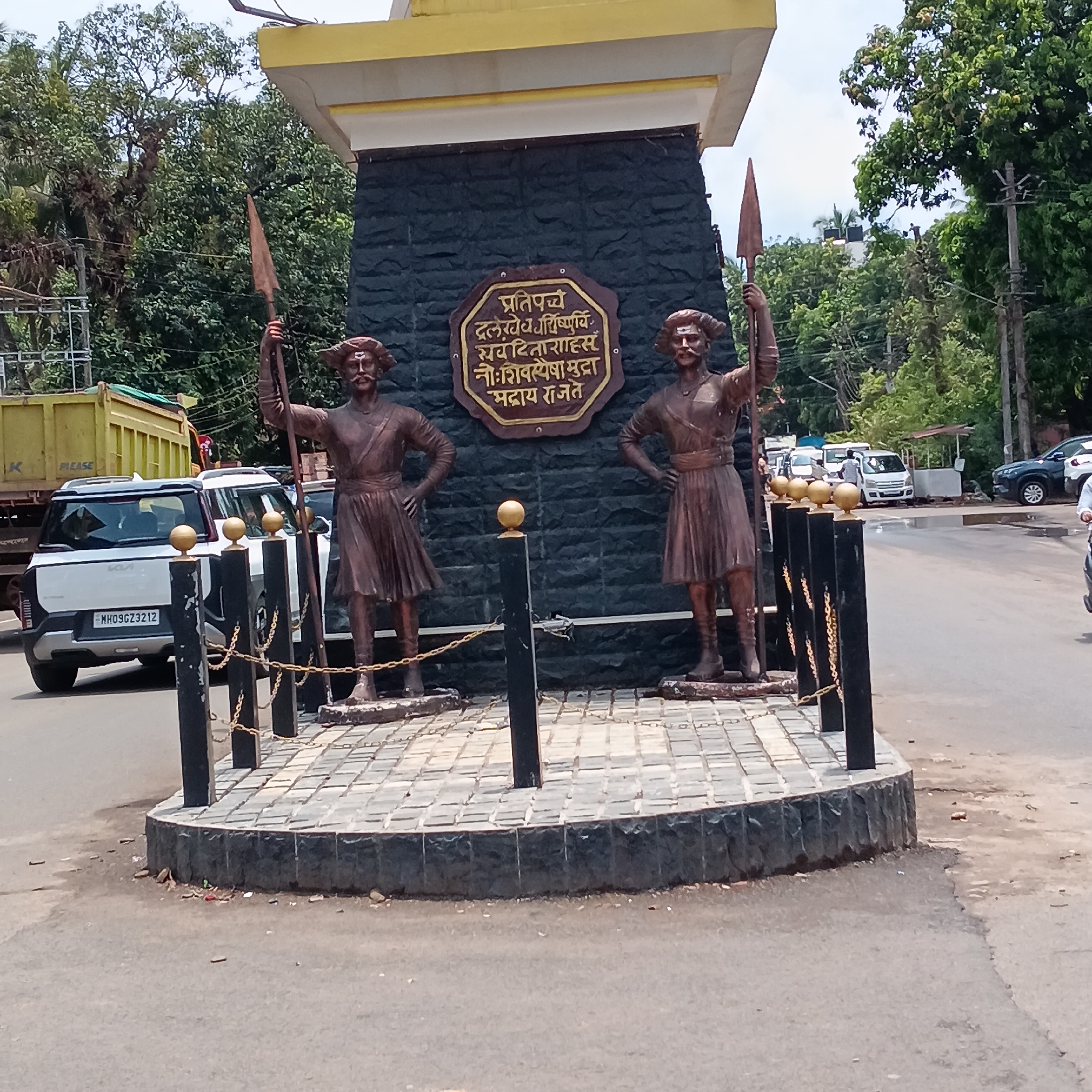 The image features a roundabout in Oxel, India, showcasing two bronze statues of warriors. The statues stand on a raised platform made of gray pavement stones, bordered by black stone. A black structure behind the statues has a bronze plaque with script in an Indian language. Short black posts, topped with gold-colored spheres, encircle the statues, linked by chains.
The scene appears to be an urban street intersection. Behind the roundabout, a yellow dump truck and other vehicles are visible. Green trees form a backdrop, and a gray sky suggests overcast weather. The overall scene depicts a public monument located within a busy street environment.