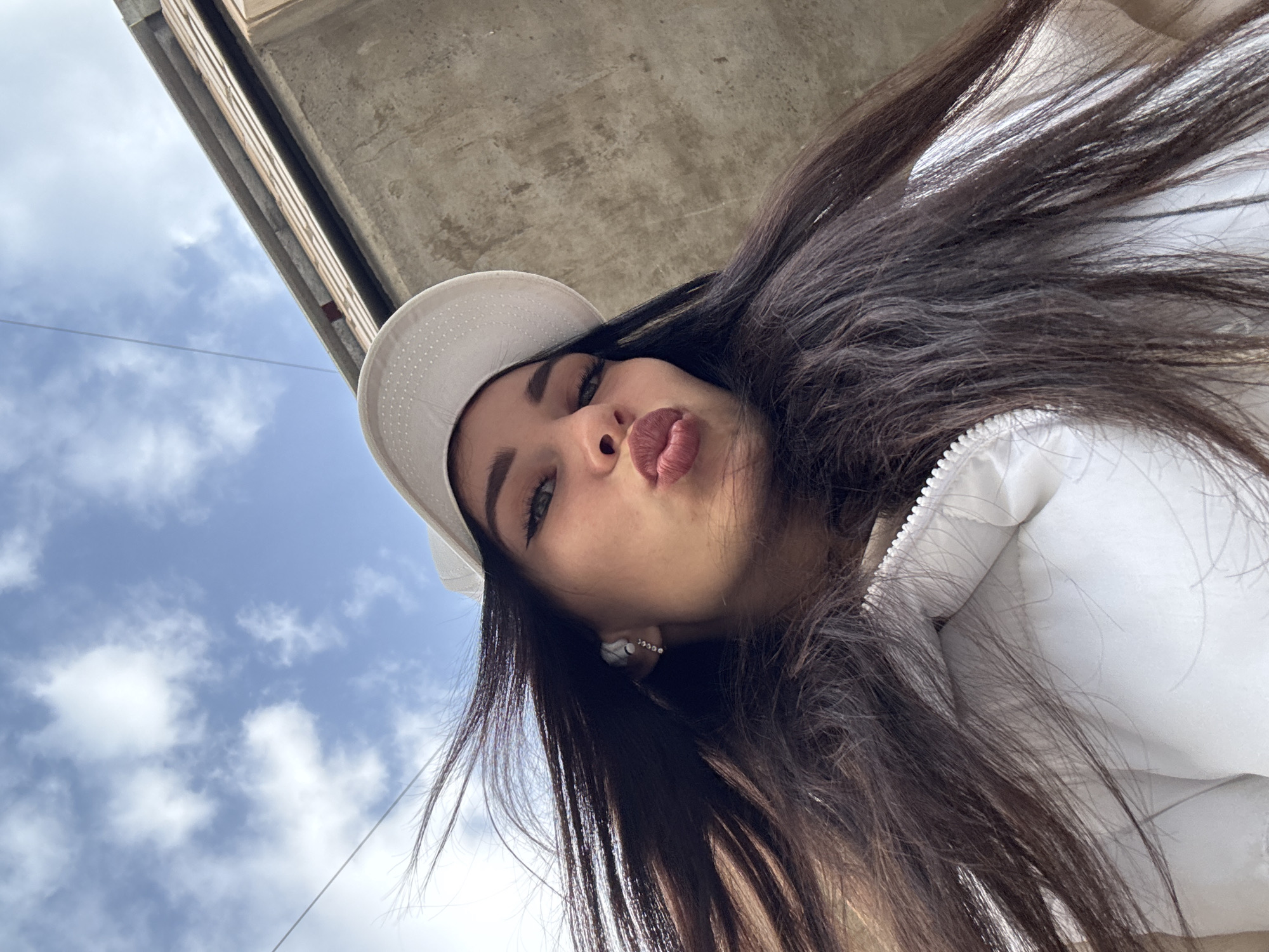 A woman with dark hair and wearing a white hat and jacket is looking down at the camera. She is puckering her lips and appears to be taking a selfie. Above her is a grey concrete structure and a view of a partly cloudy sky. Based on the location requested, it is likely that the image was taken in the city of Chaikovsky, Russia.