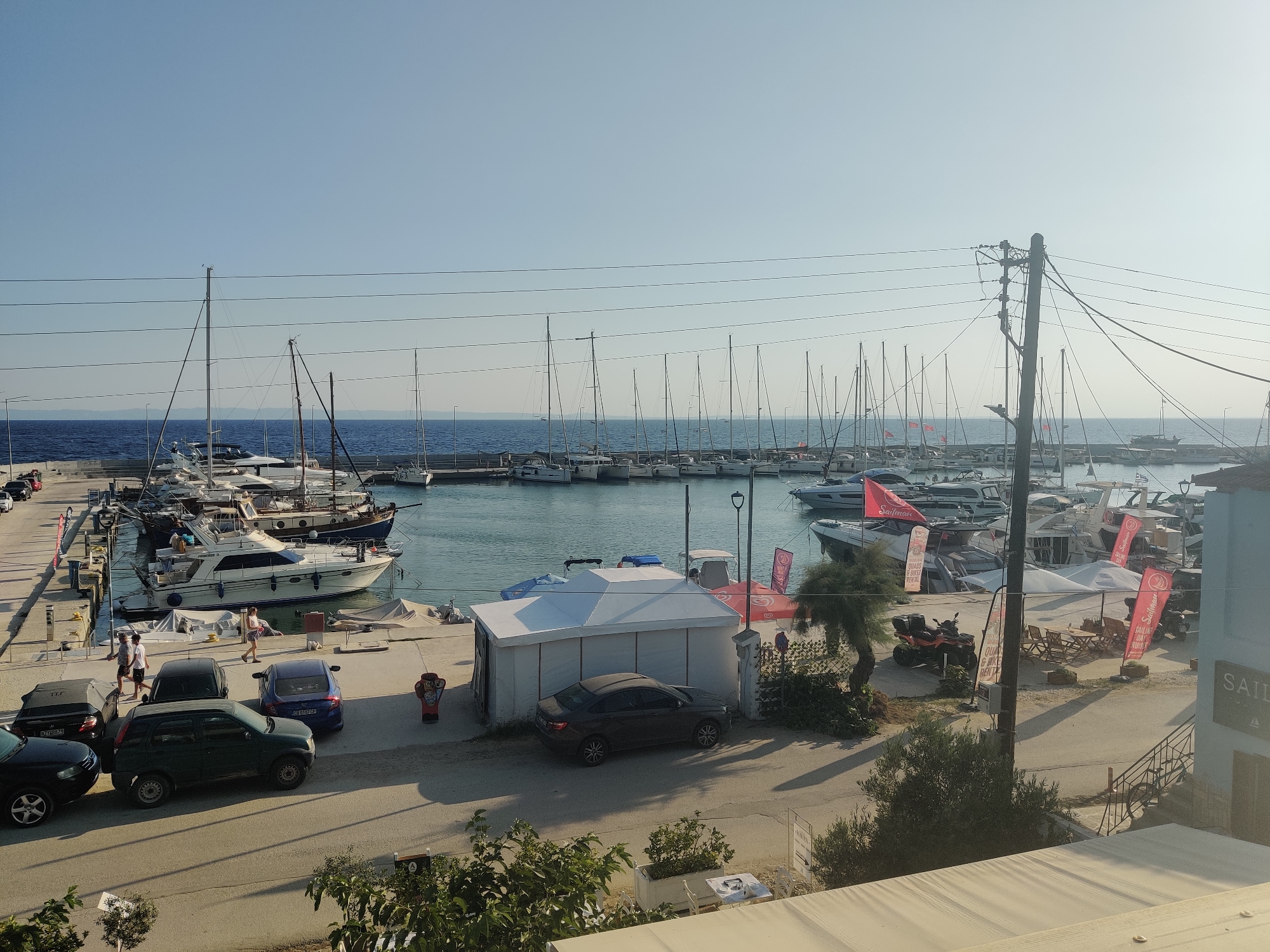 An outdoor, elevated view shows a harbor in Sithonia Municipal Unit, Greece, with numerous sailboats and motor yachts moored in the calm blue water. The harbor is lined with a paved walkway and a road. Several cars are parked along the road and walkway. On the walkway, there are white tents, outdoor seating areas, and some people walking. Red flags with white lettering are visible on some of the boats and a tall pole. The sky is clear and blue.