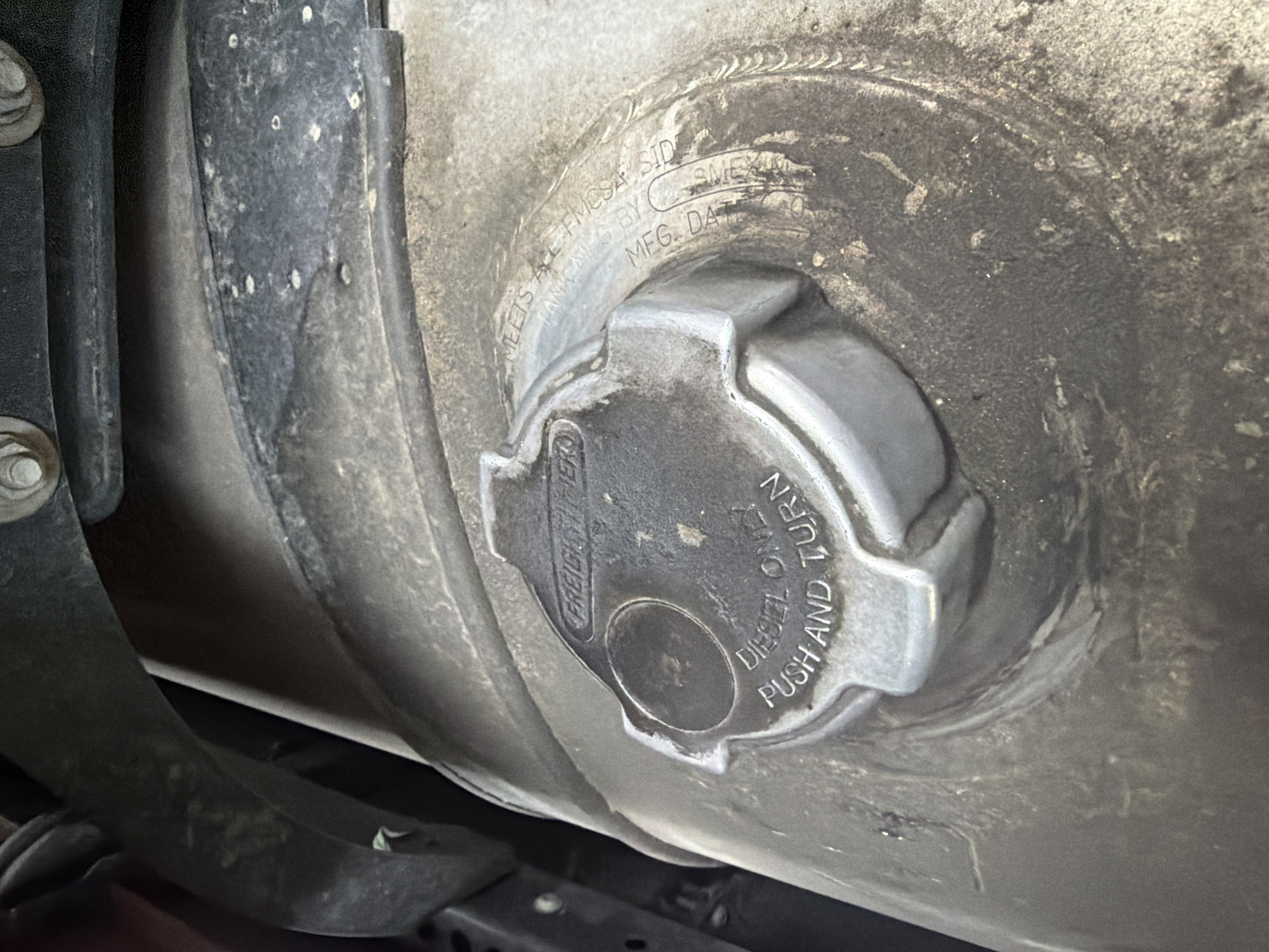 A close-up, eye-level shot captures a dusty, metal diesel fuel cap on a truck. The cap, featuring the "FREIGHTLINER" logo and instructions to "DIESEL ONLY PUSH AND TURN," is set against a grimy, cylindrical fuel tank. Faint inscriptions on the cap read, "MEETS ALL FMCSA & SID TANK MLS BY SWE," and "MFG. DA". The surrounding area is obscured by dark, shadowy components, likely part of the truck's undercarriage. The scene suggests a maintenance or operational context for a commercial vehicle.